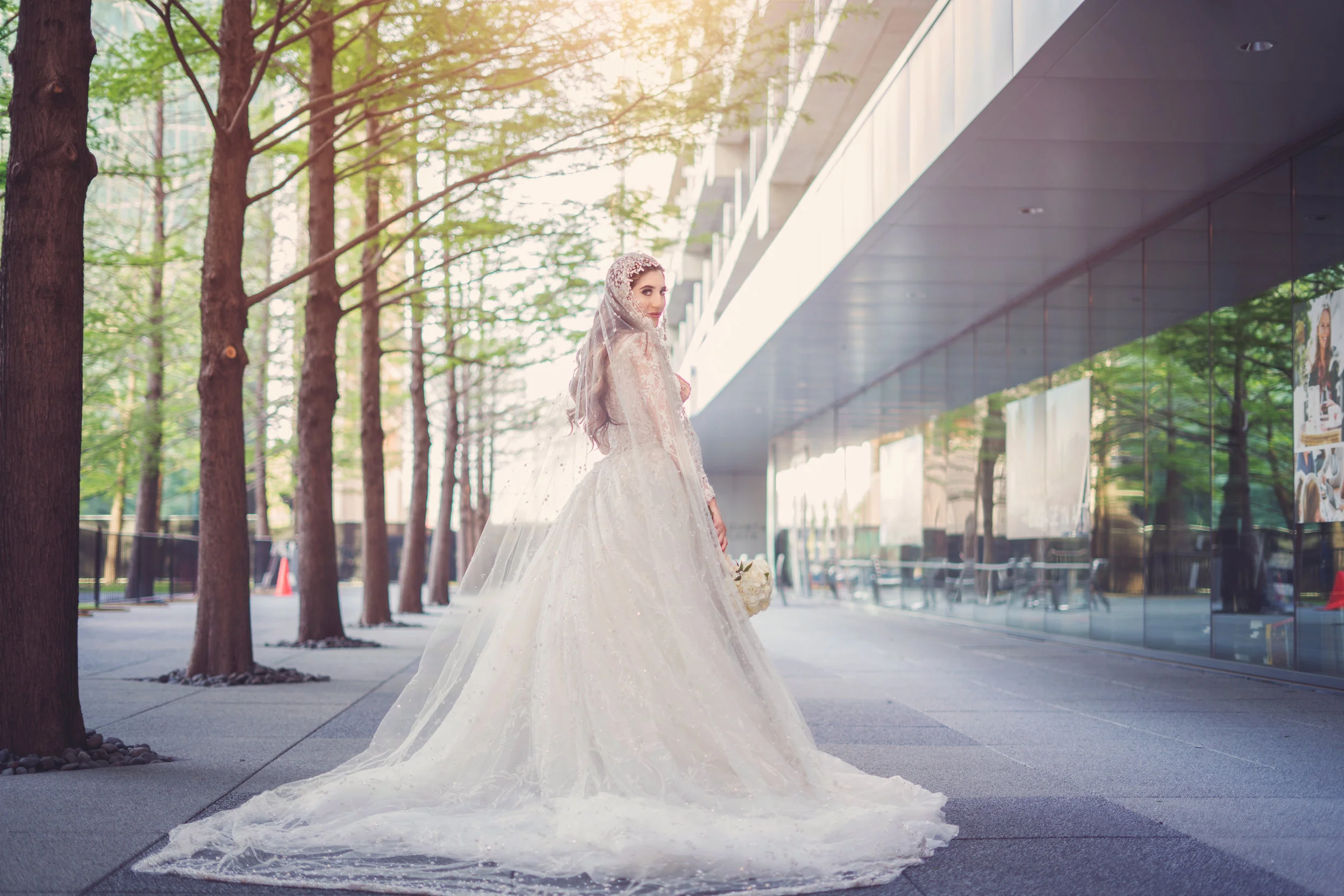 Bride in lace wedding gown walking outdoors near modern building, surrounded by trees.