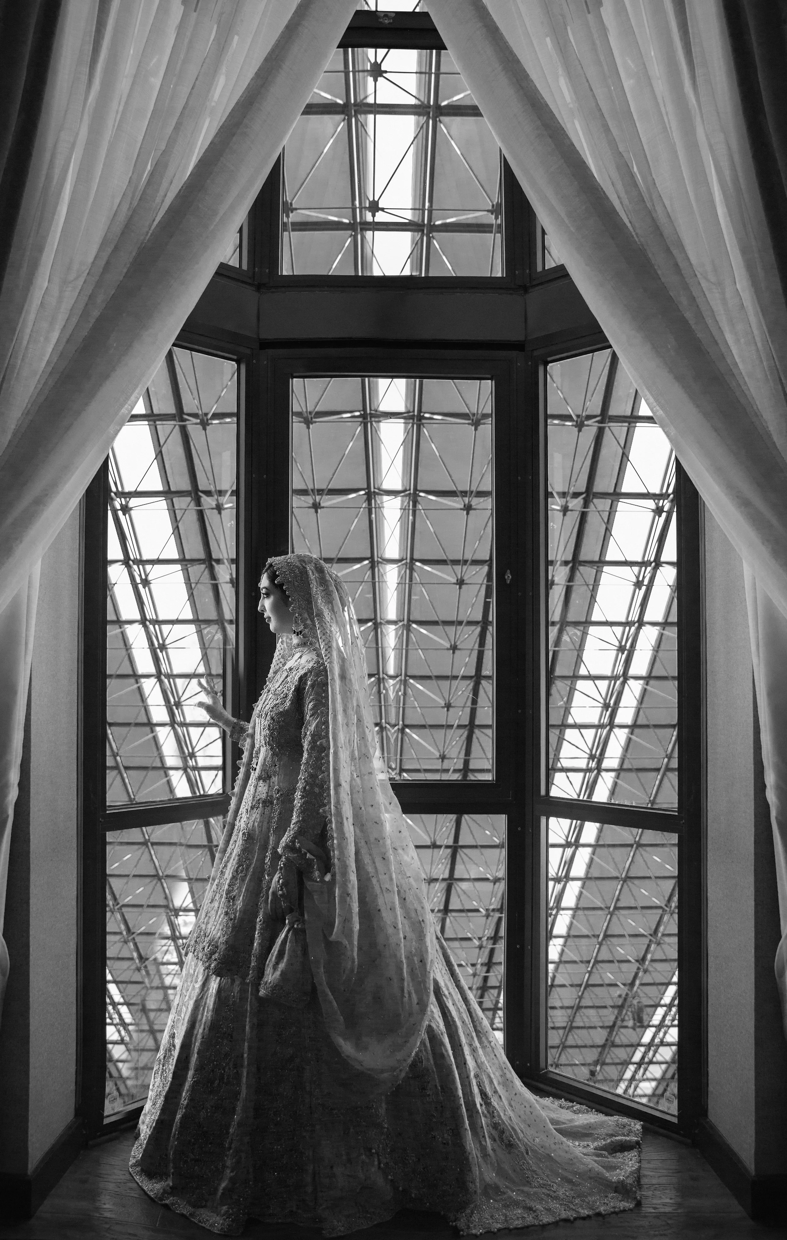 Black and white photo of a bride in an elaborate gown and veil standing by a large window with curtains, looking out.