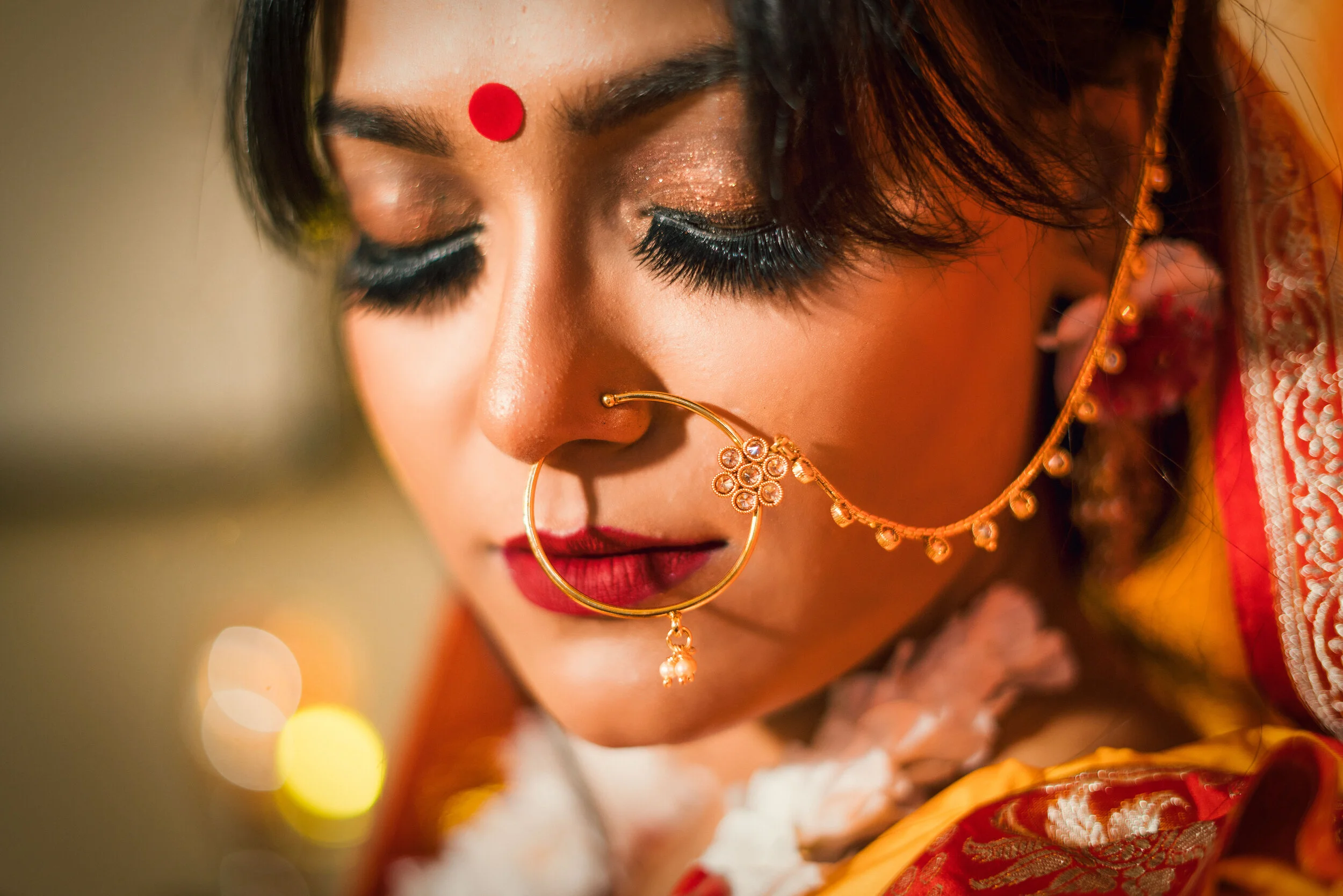 Close-up of a woman with traditional Indian jewelry, wearing a nose ring with chain and a bindi, adorned in a red and gold sari.