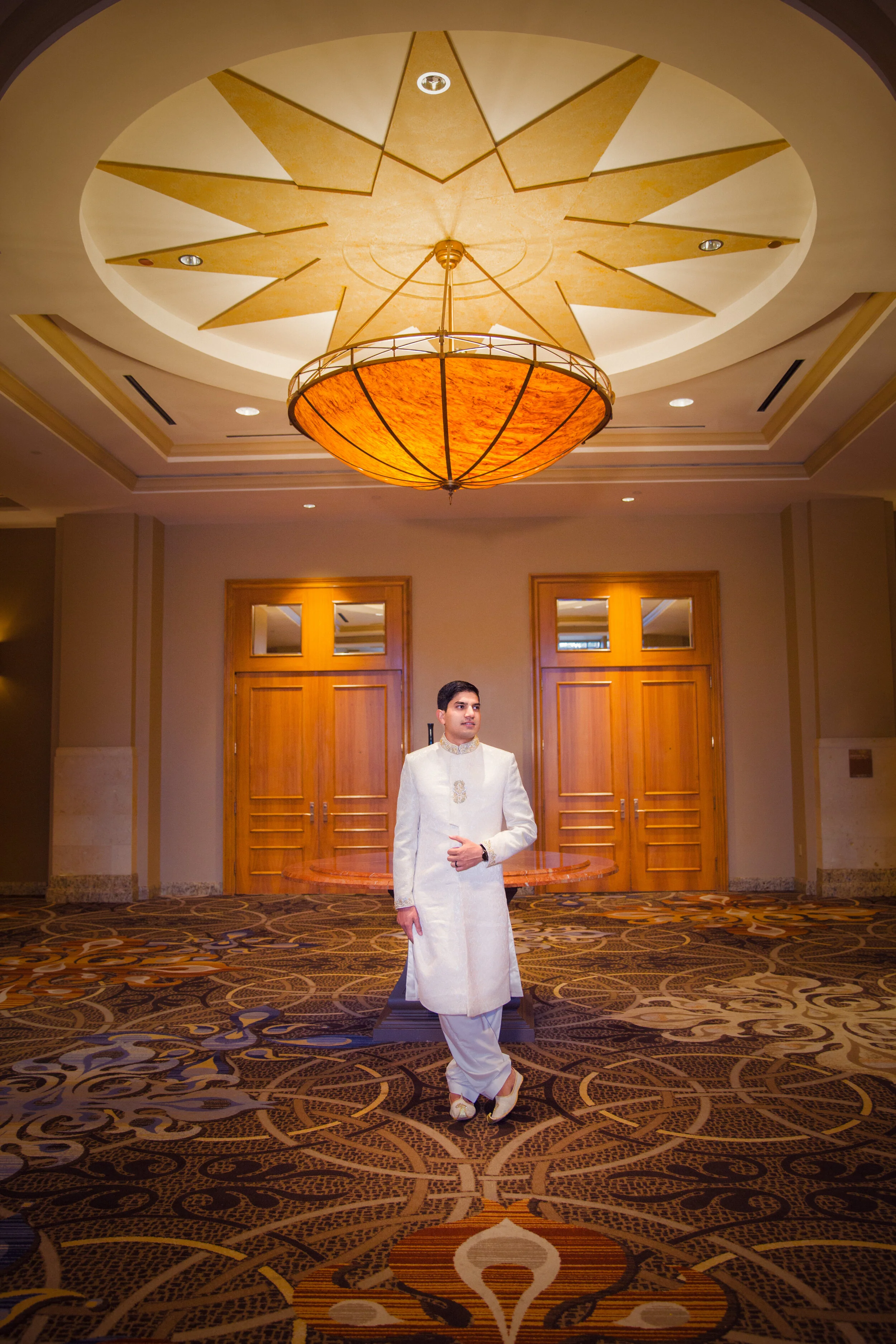 Man in traditional white attire standing in an elegant room with ornate ceiling and wooden doors.