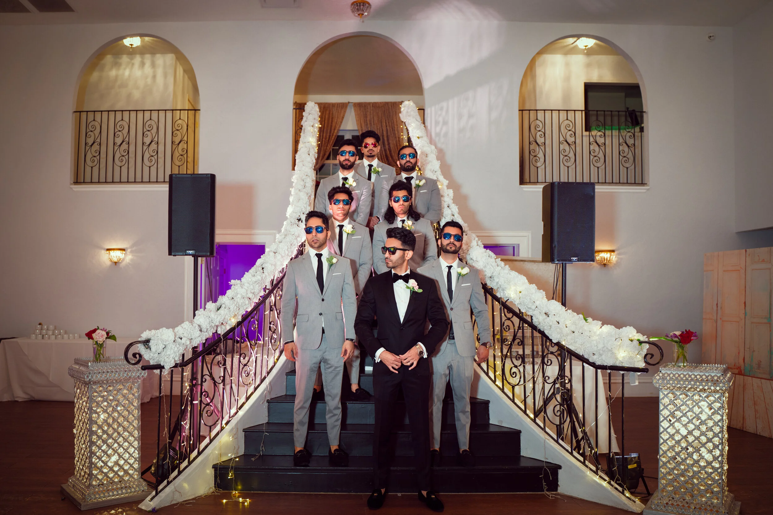 A group of groomsmen wearing sunglasses stand on a decorated staircase, with one in a black tuxedo and the others in gray suits.
