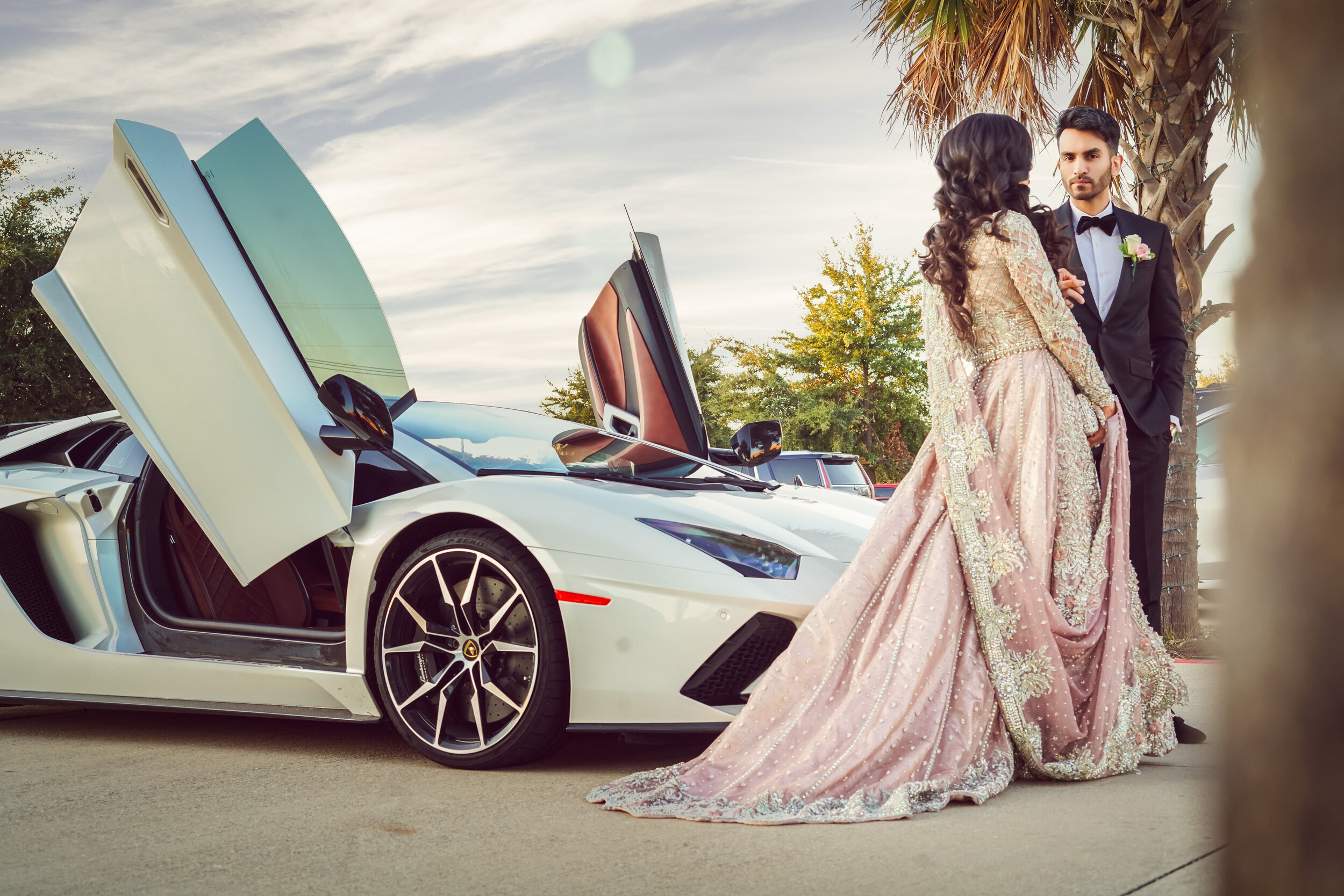 A couple dressed in formal attire stands next to a luxurious white sports car with open vertical doors, under a palm tree, with a scenic backdrop.