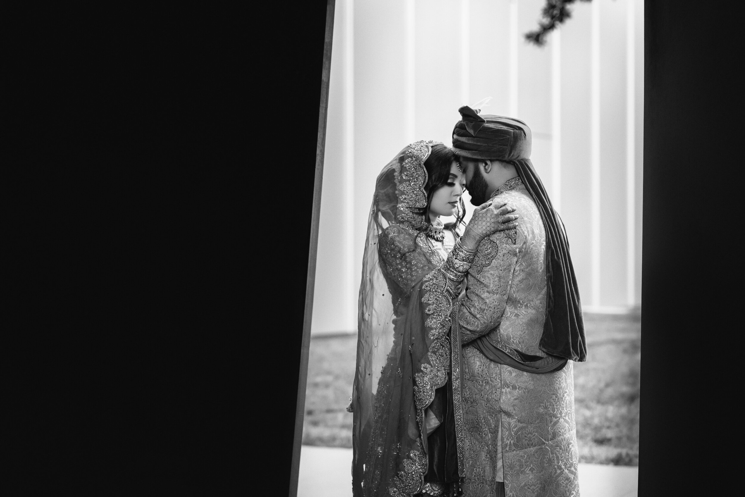 A bride and groom in traditional attire, embracing closely in a black and white photograph. The bride is wearing ornate jewelry and a veil, while the groom has a decorative headscarf. The setting appears intimate and romantic.