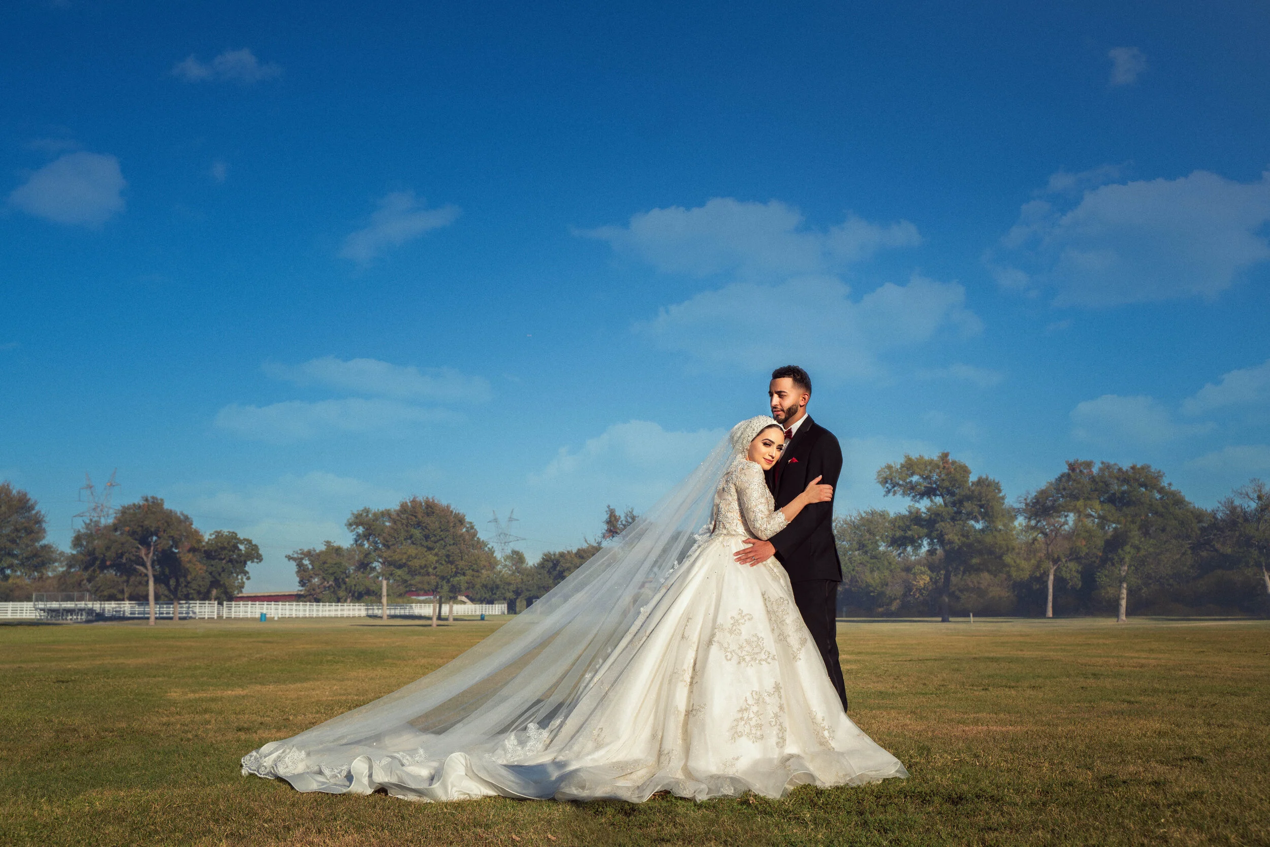 Bride and groom embracing on a grassy field with a clear blue sky.