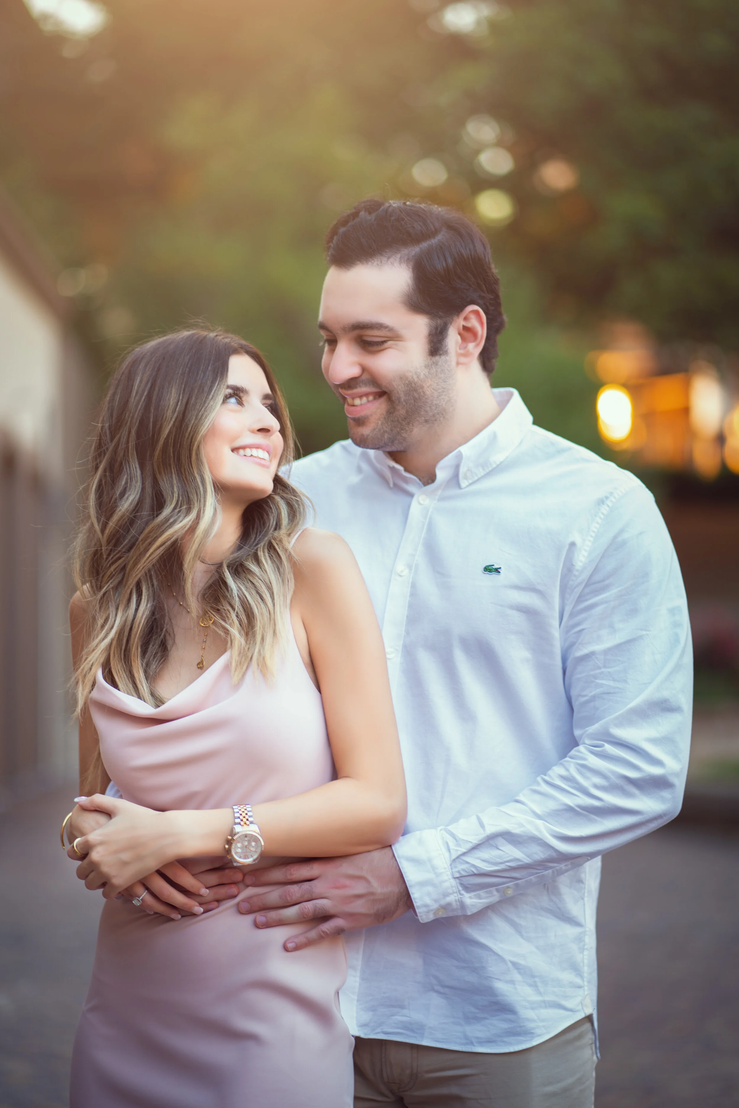 A smiling couple embracing outdoors in soft natural light, with the woman wearing a pink dress and watch, and the man in a white shirt, surrounded by a blurred green background.