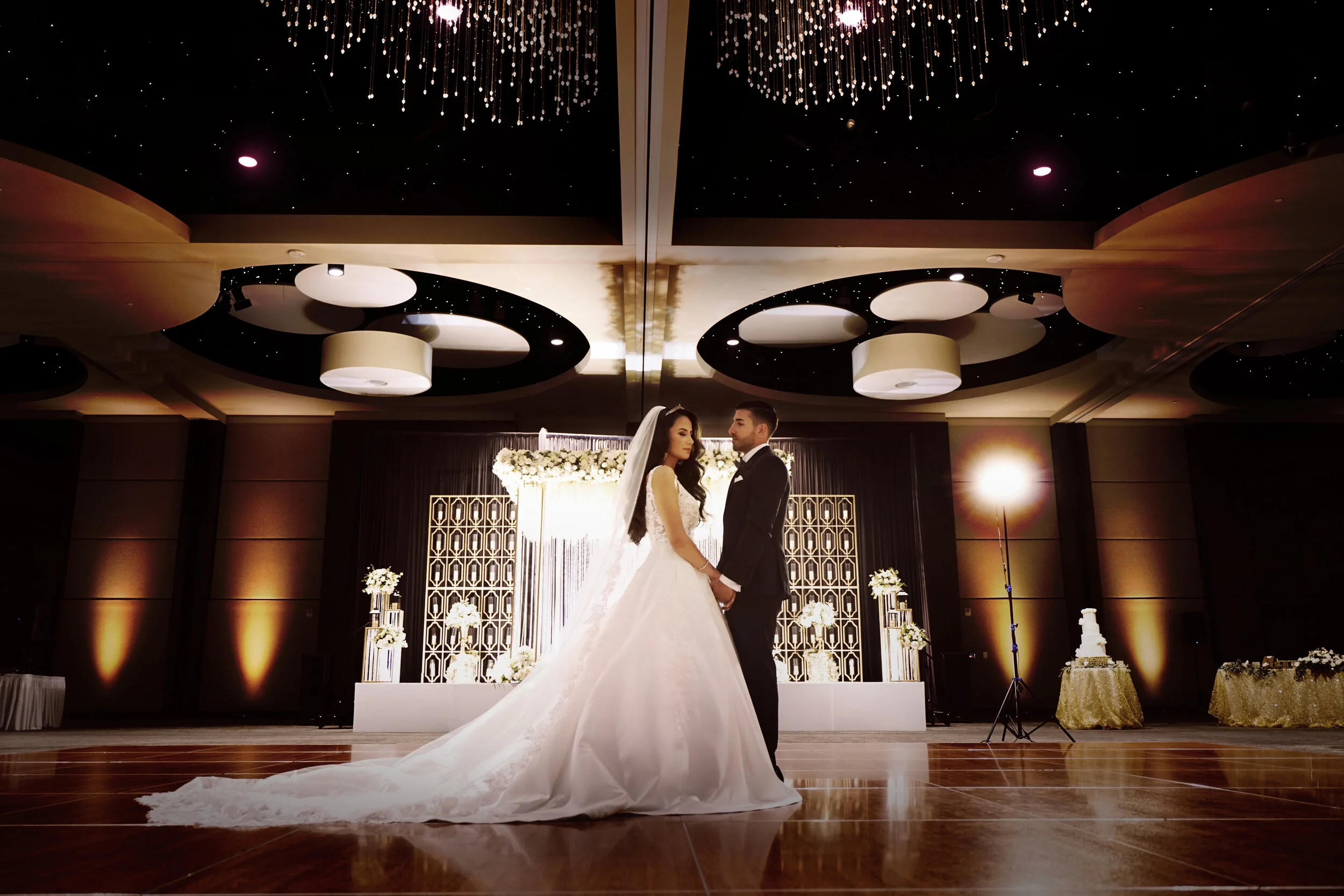Bride and groom standing in a decorated wedding venue with elegant chandeliers and floral arrangements.