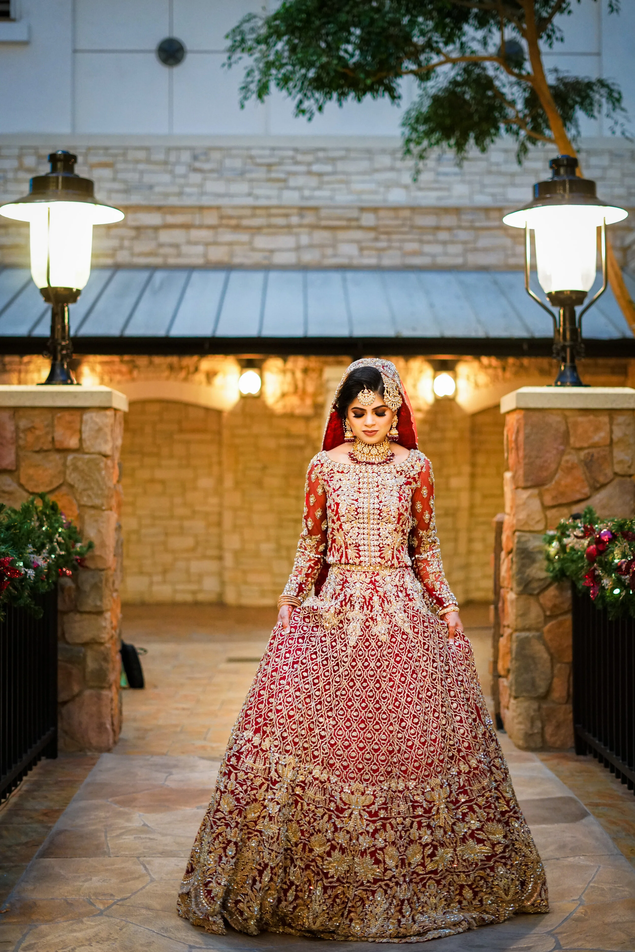 Bride in red and gold traditional dress with intricate designs, standing in an outdoor setting with stone walls and lanterns.