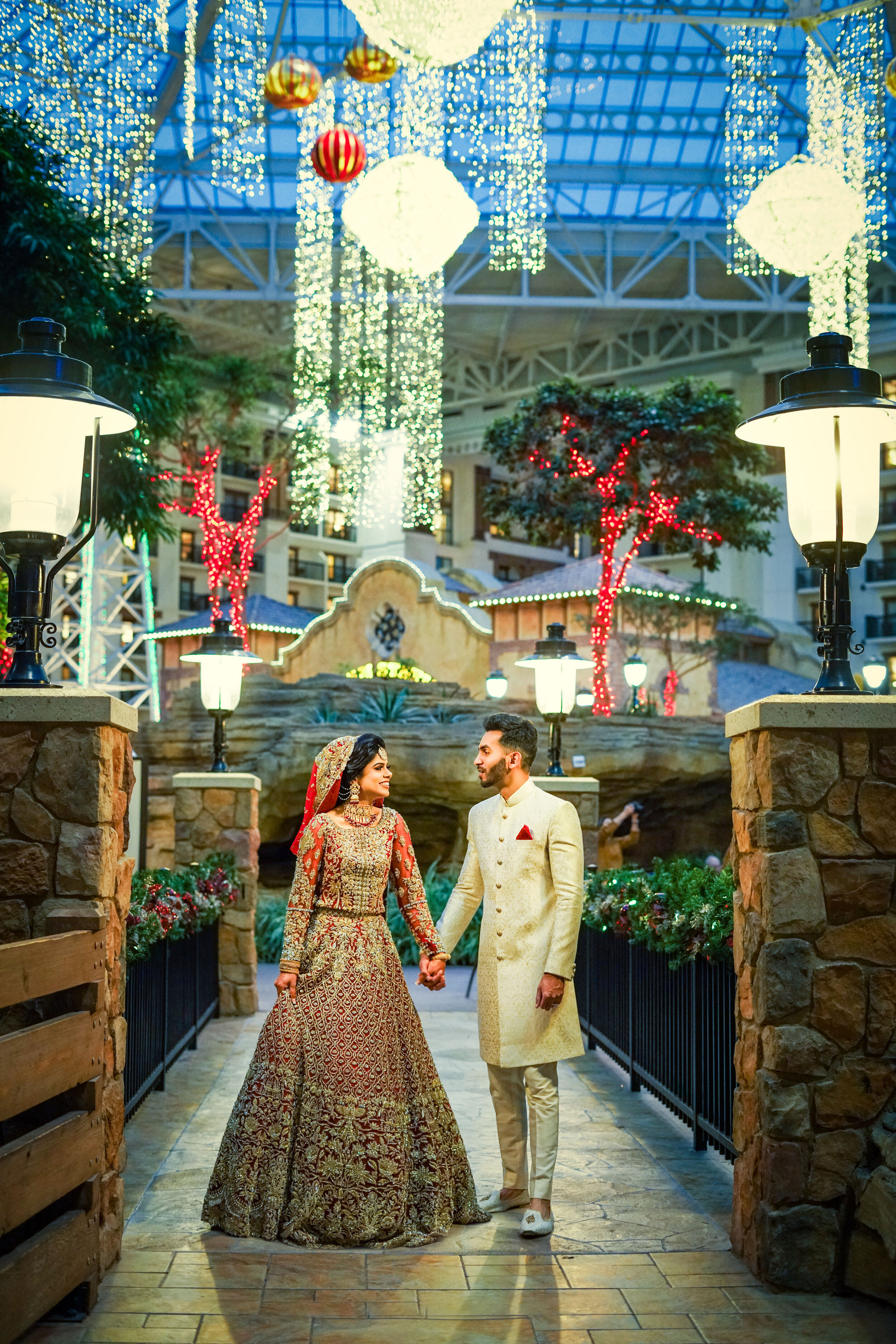 A couple in traditional South Asian wedding attire stands hand in hand under decorative lights in an indoor garden with stone walls and festive decorations.