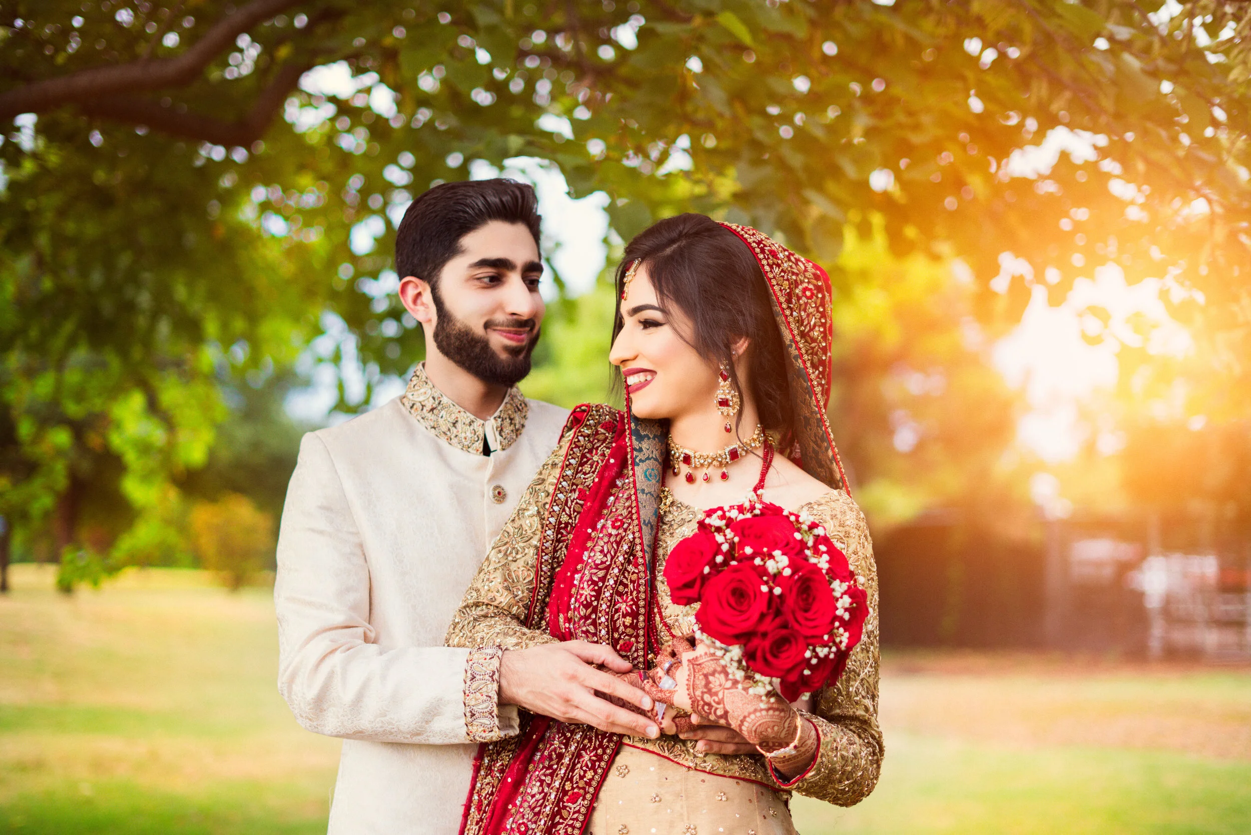 A couple dressed in traditional South Asian wedding attire stands outdoors, surrounded by greenery. The bride is holding a bouquet of red roses and wears an ornate red and gold outfit. The groom is in a cream-colored sherwani, smiling at the bride.