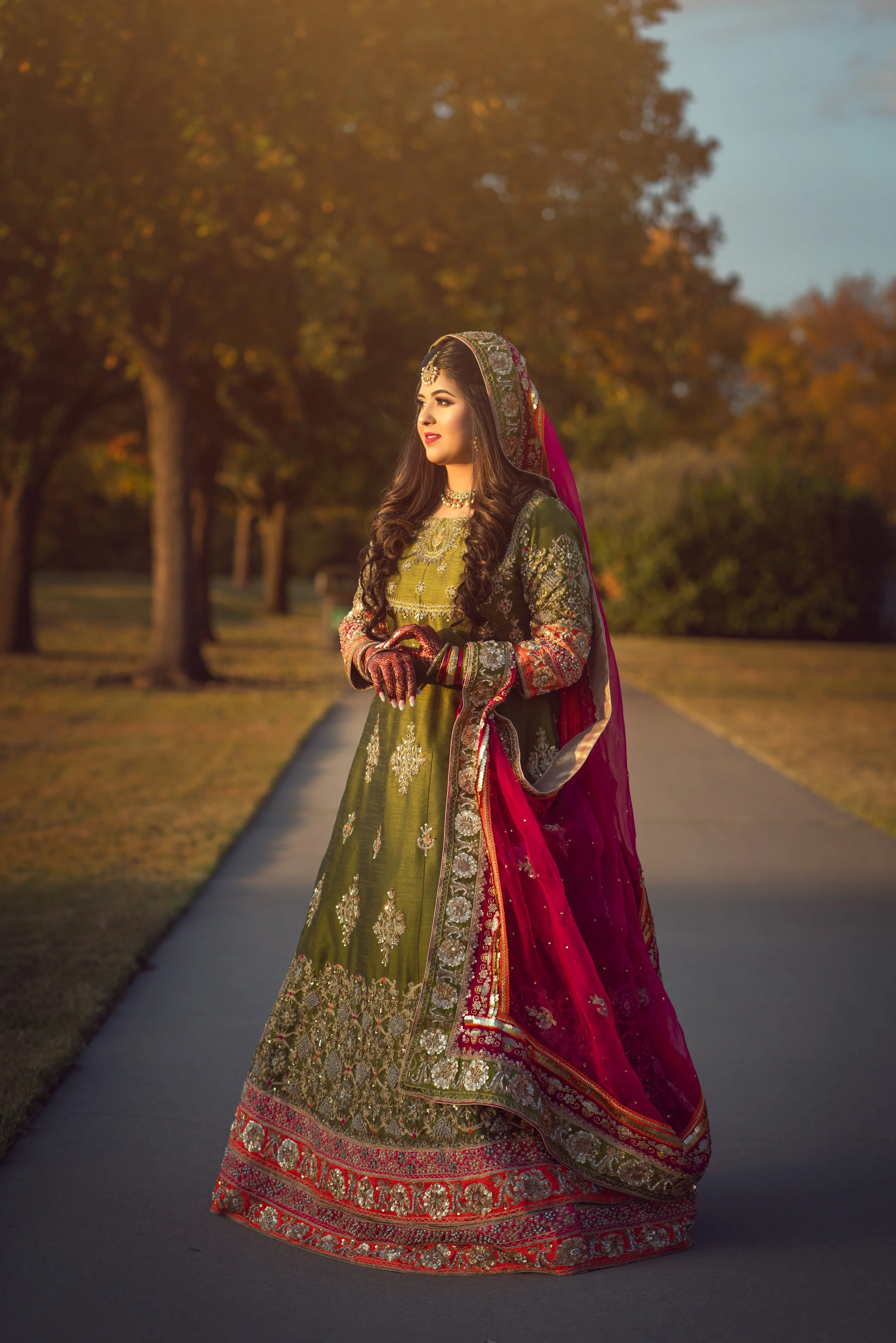 Woman in traditional South Asian bridal attire with green and pink embellished dress, standing outdoors on a path lined with trees.
