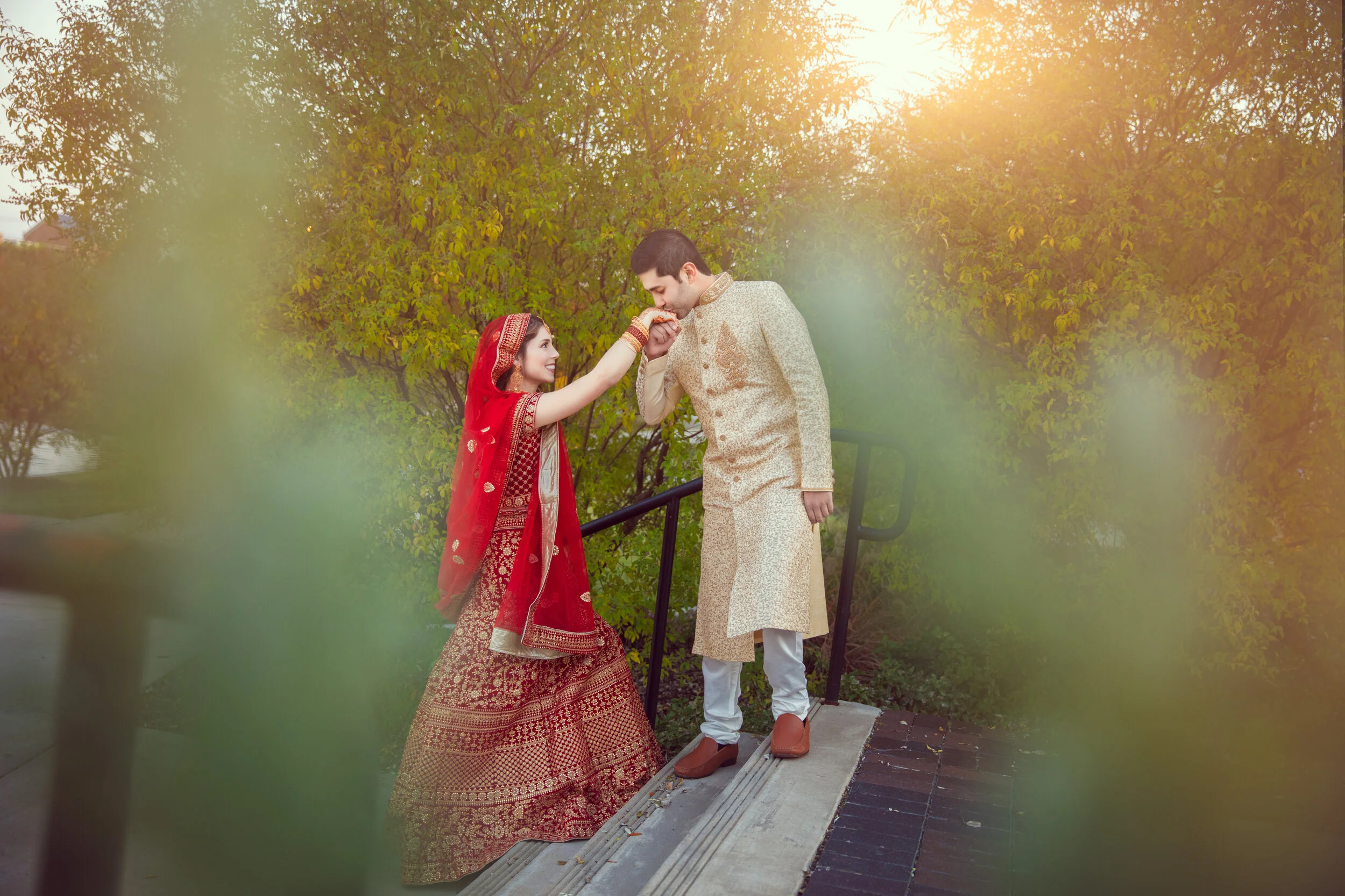 A couple in traditional South Asian wedding attire, with the bride wearing a red lehenga and the groom in a beige sherwani, outdoors with greenery and soft sunlight.