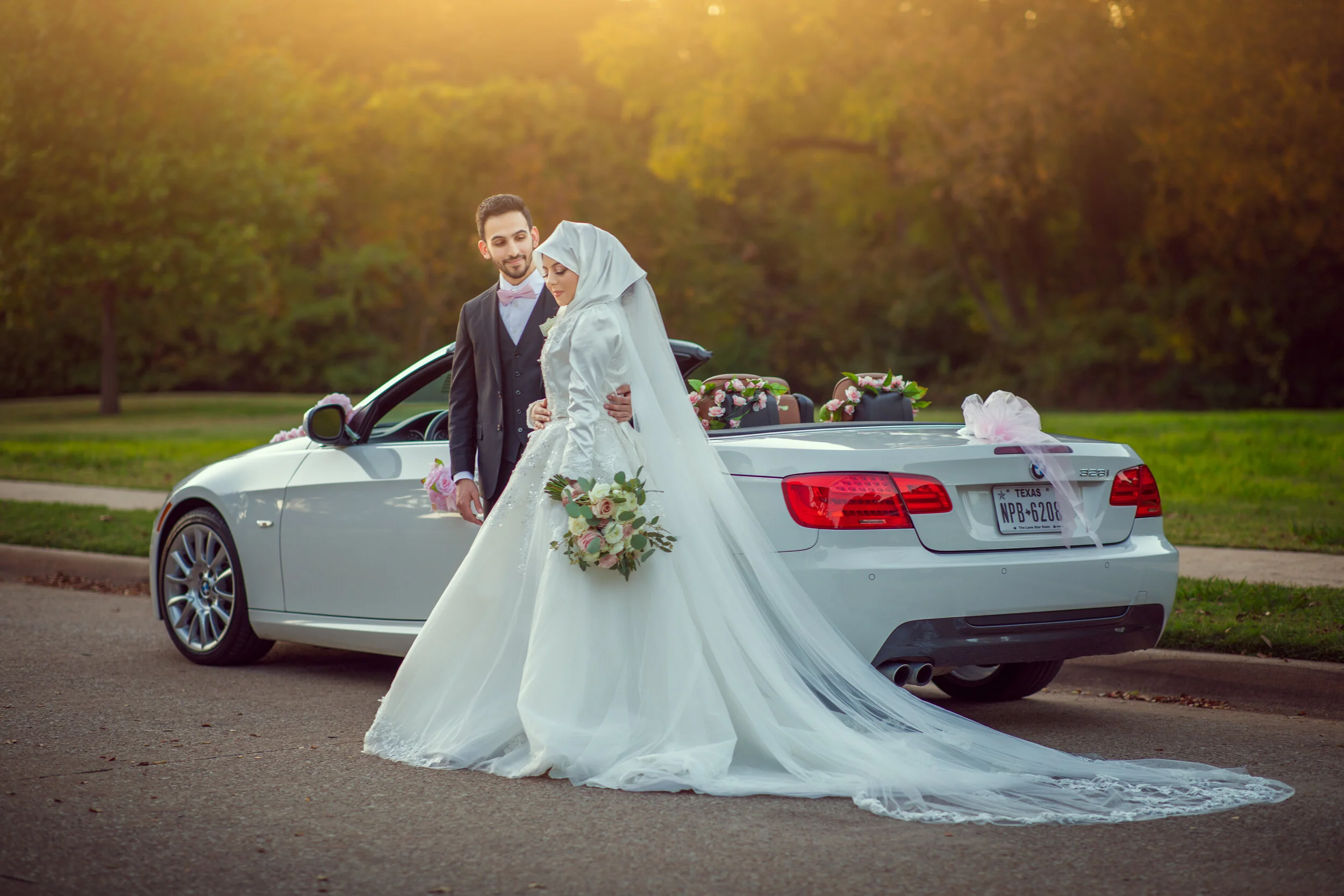 Bride and groom standing near a decorated white convertible car in a scenic outdoor setting; bride wearing a white gown and holding a bouquet.