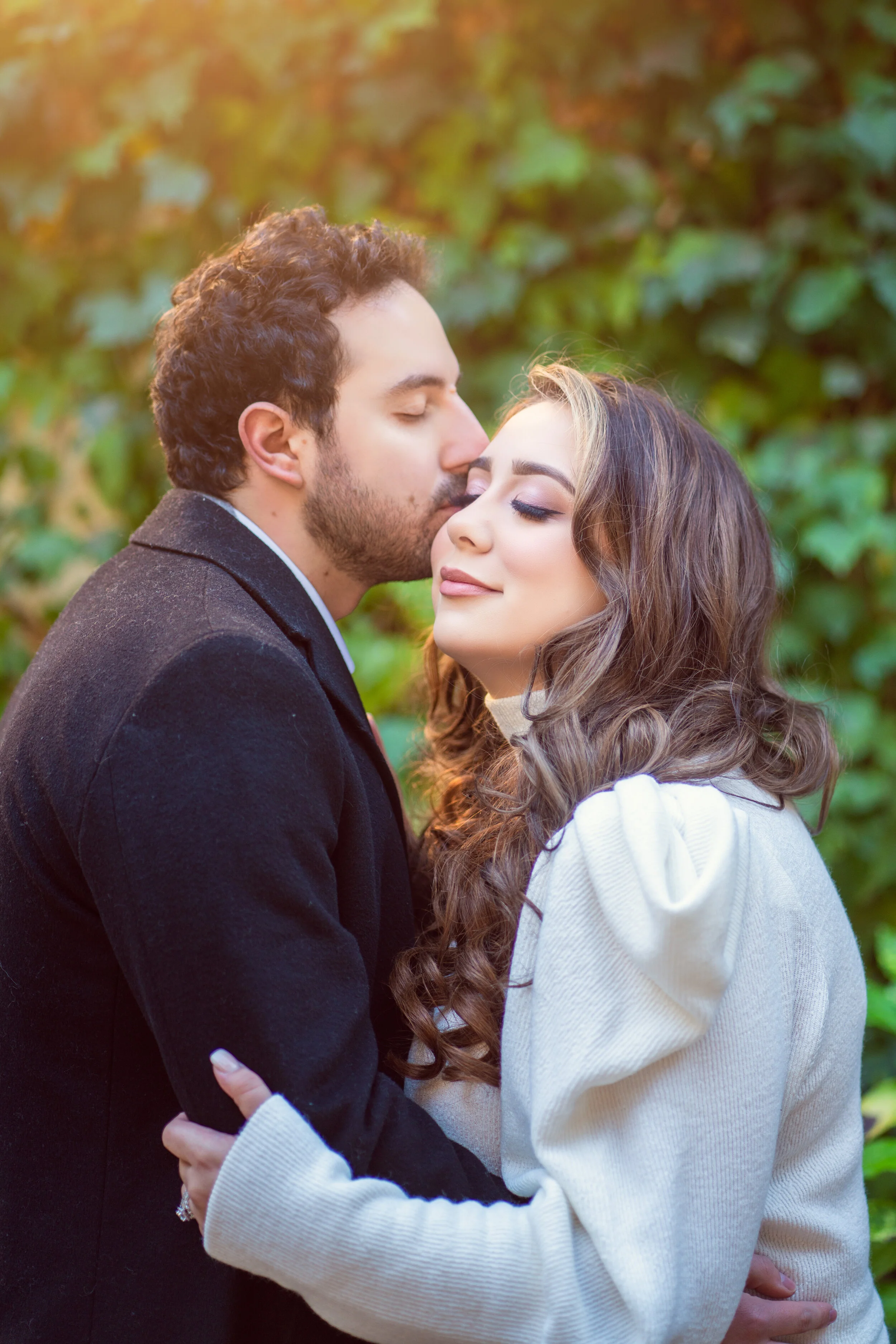 A couple embraces lovingly in front of a leafy green background. The man, wearing a black coat, kisses the forehead of the woman, who is dressed in a white sweater. Both appear content and relaxed.