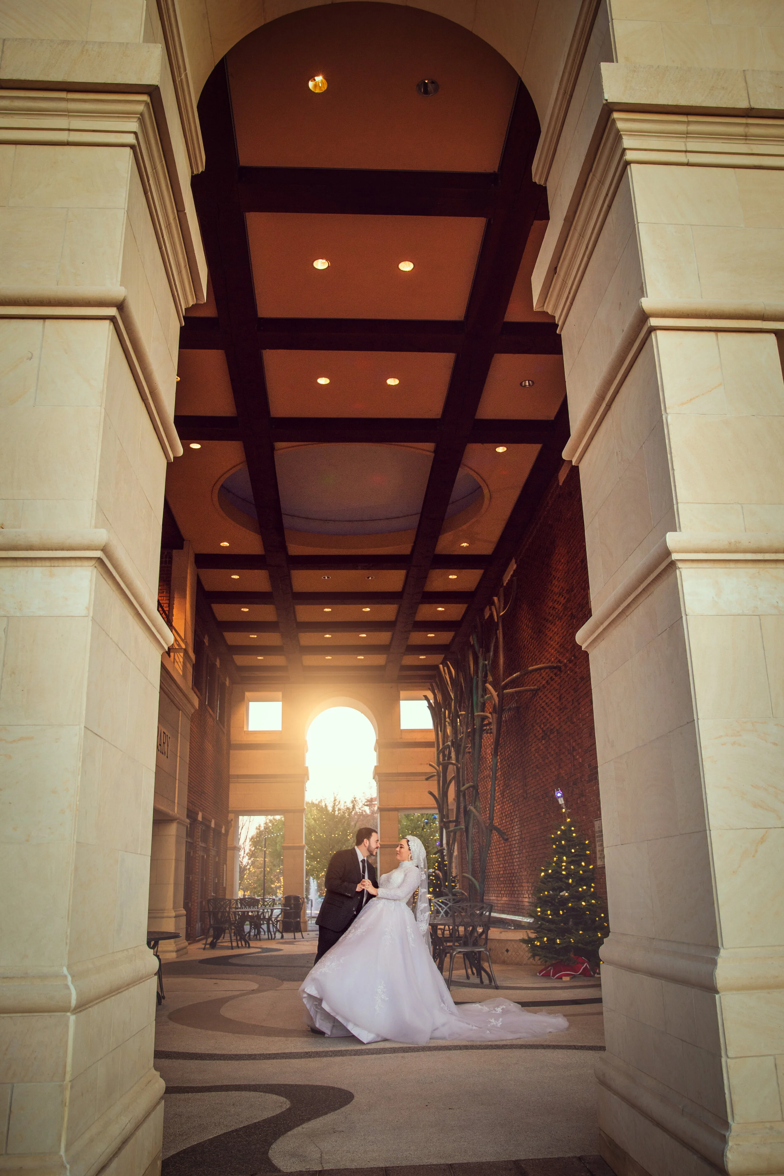 A bride and groom dancing under a grand archway, with a Christmas tree in the background and soft sunlight streaming through the entrance.