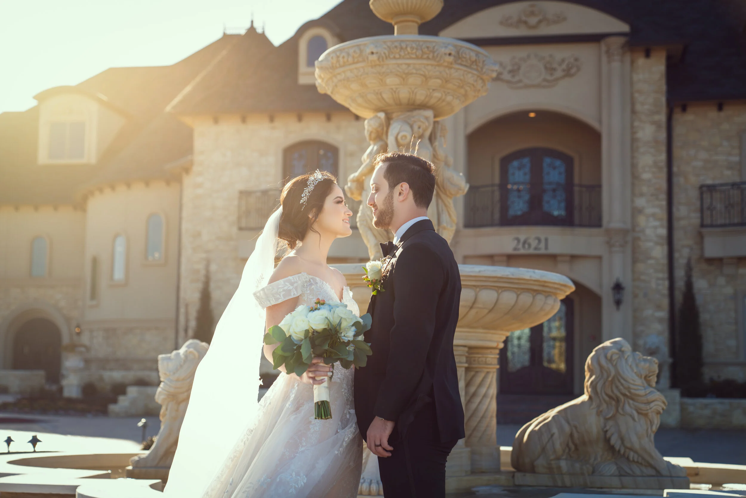 Bride and groom standing in front of a large stone fountain with a mansion in the background. The bride is wearing a white wedding gown and holding a bouquet of white flowers, while the groom is in a black suit.