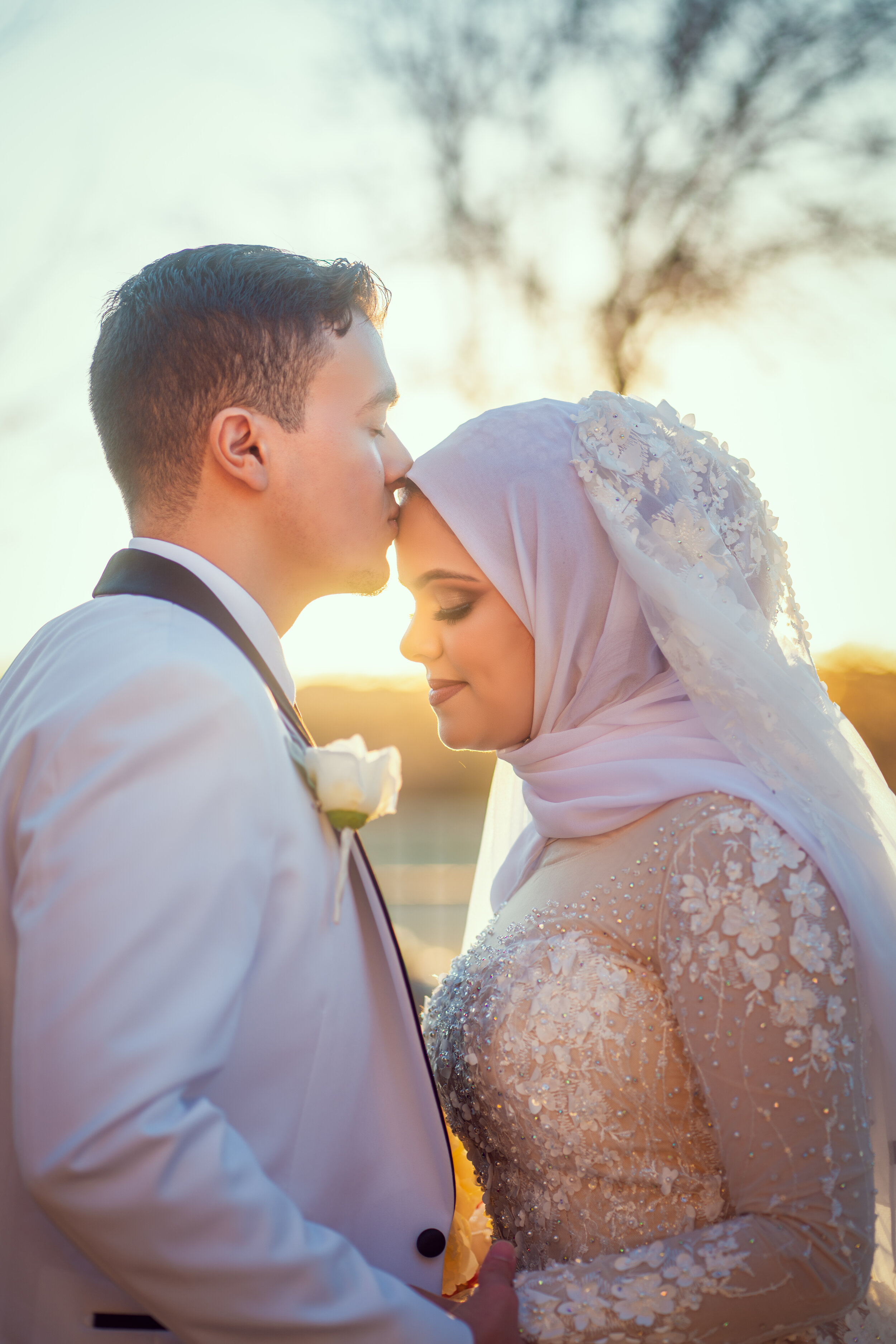 Man in white tuxedo kissing woman in bridal gown on forehead outdoors at sunset.