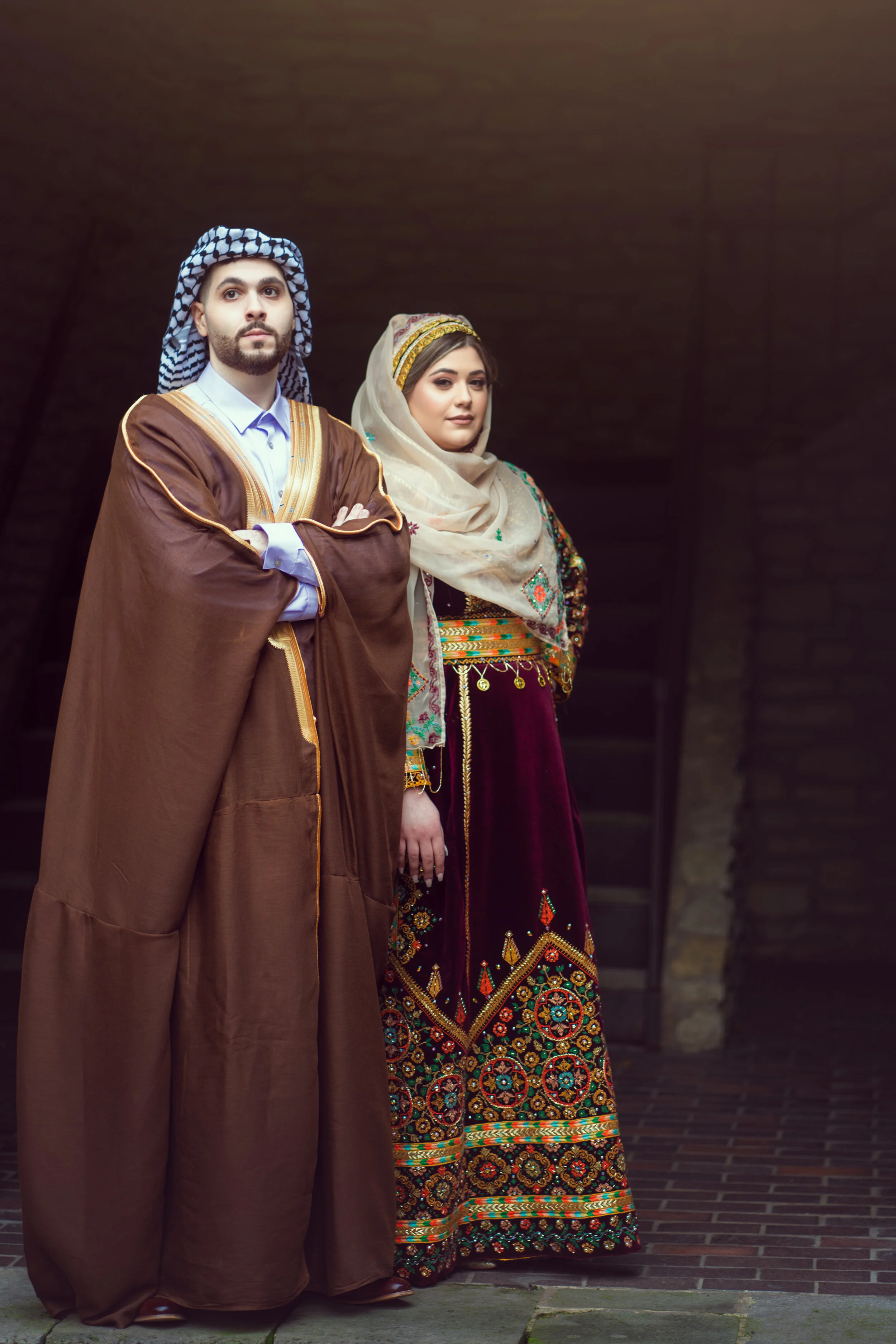 A man and a woman in traditional Middle Eastern clothing, standing in front of a dark background. The man is wearing a brown robe and checkered headscarf, and the woman is in a richly embroidered dress with a headscarf.