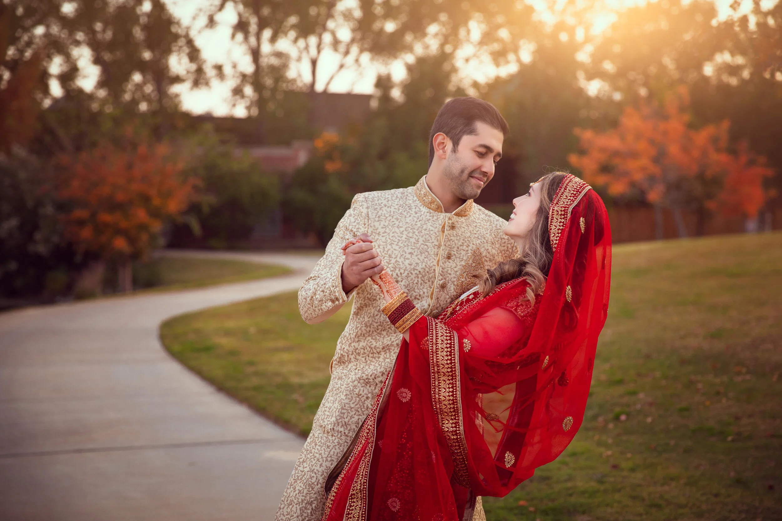 Couple in traditional attire, man in beige sherwani, woman in red saree, embracing outdoors with autumn trees and sunset in background.