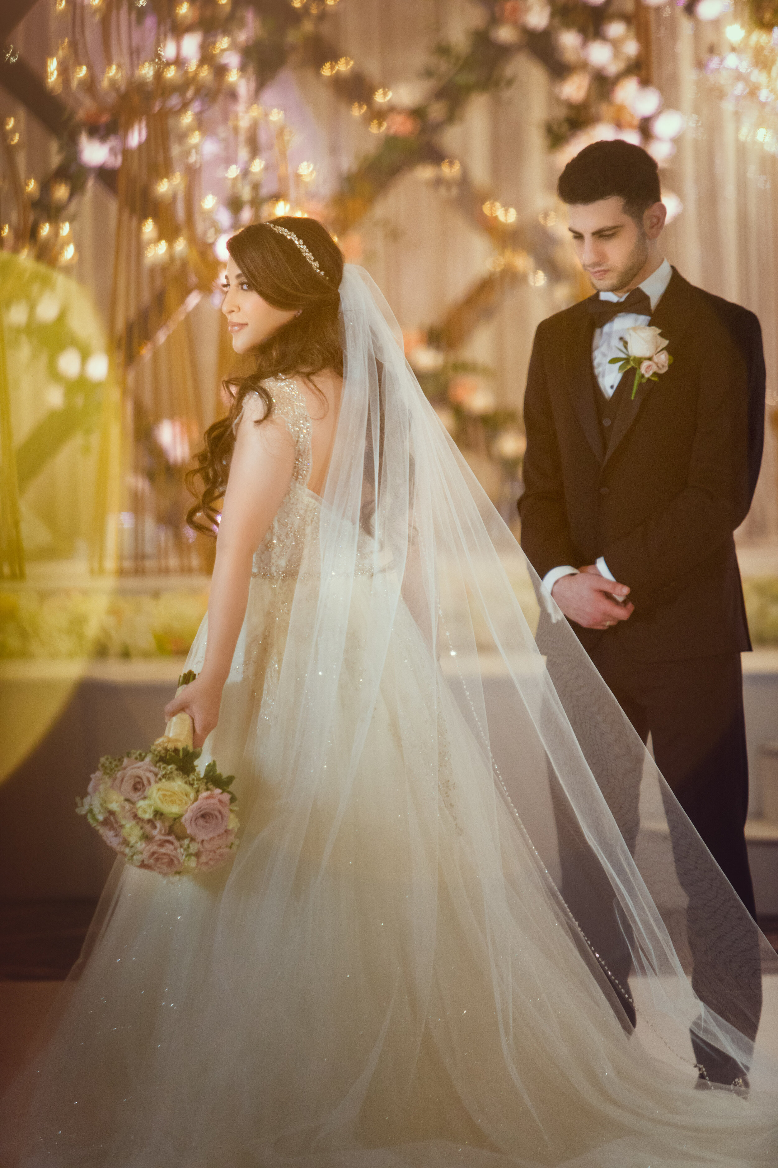 Bride and groom in formal attire, bride holding bouquet, soft lighting with floral backdrop.