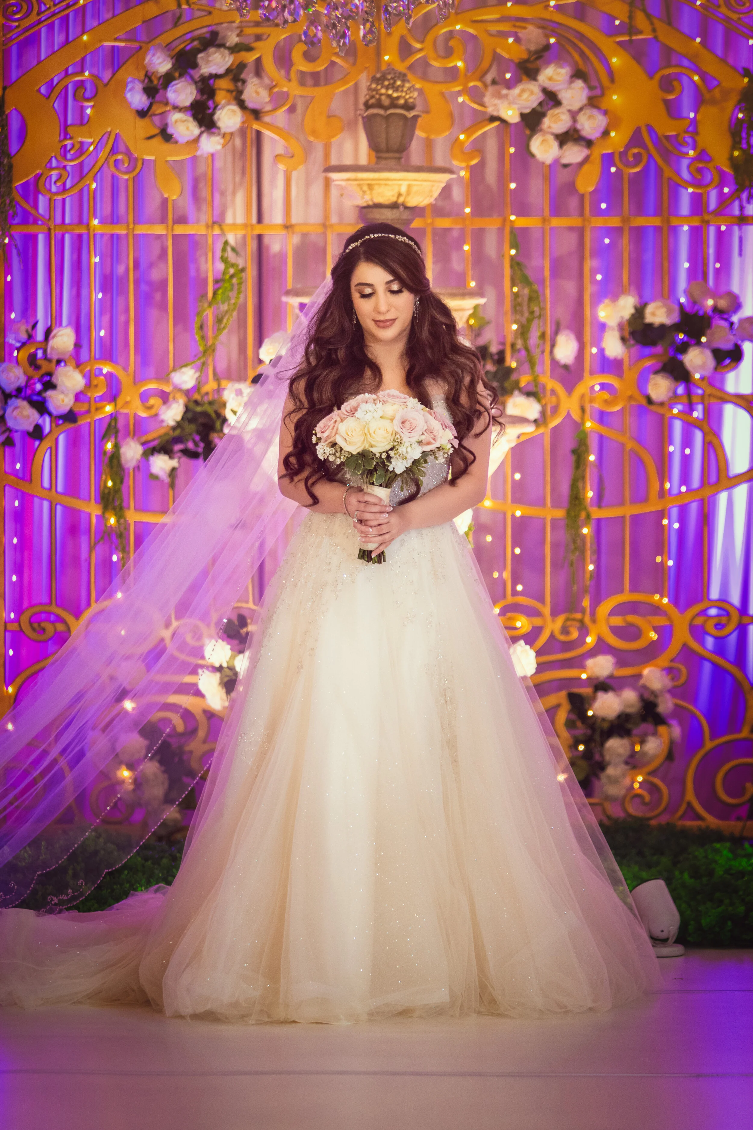 Bride in white gown holding bouquet, standing in front of ornate floral backdrop with lights.