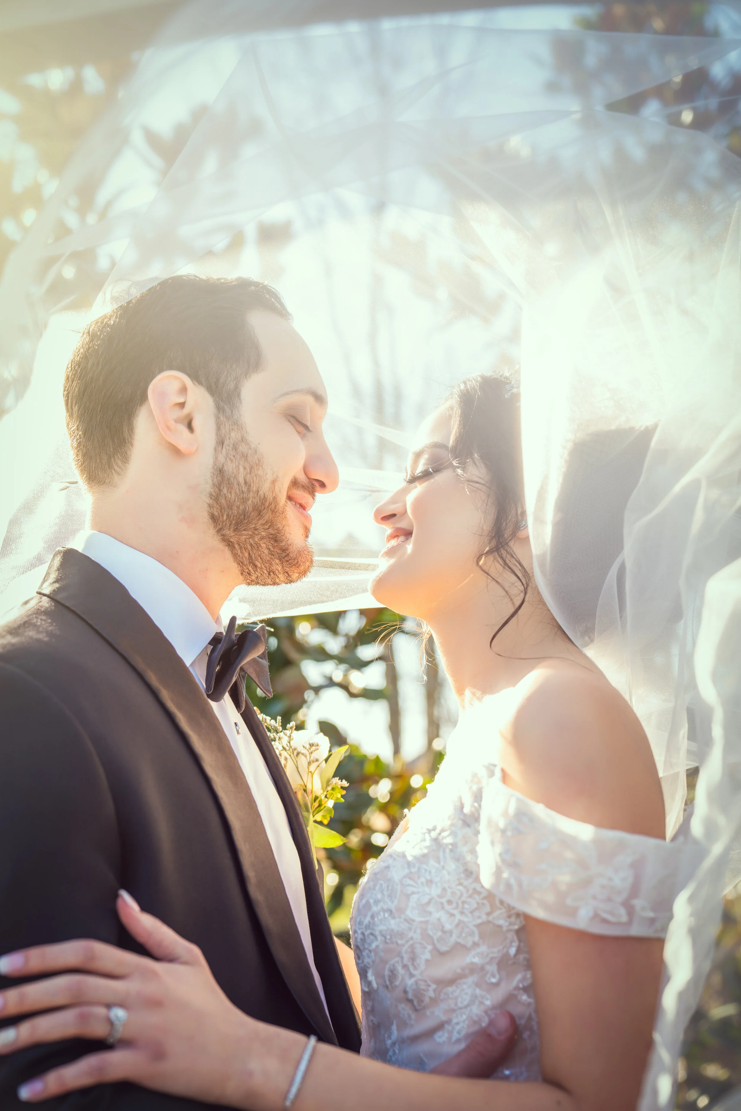 A bride and groom smiling under a veil outdoors.