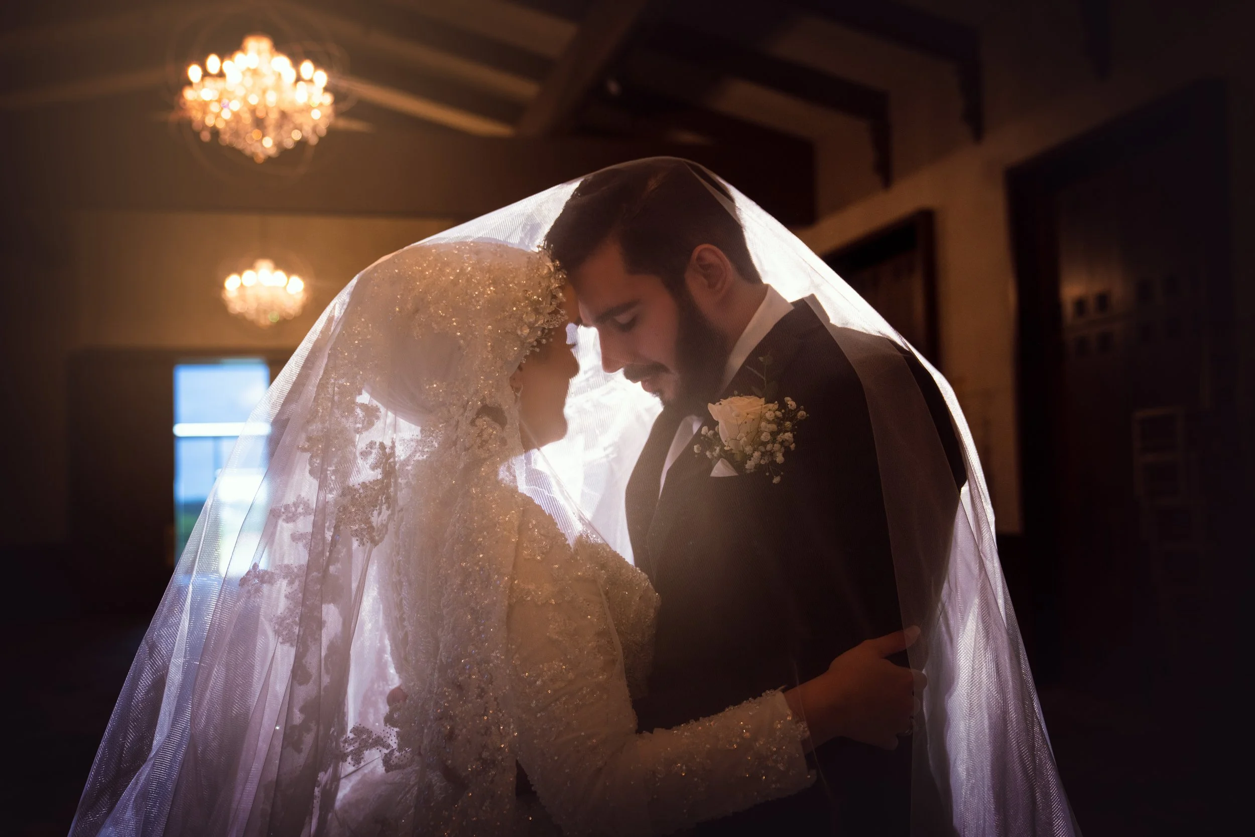 A romantic wedding scene with a bride and groom holding each other under a veil, illuminated by soft lighting from chandeliers in the background.