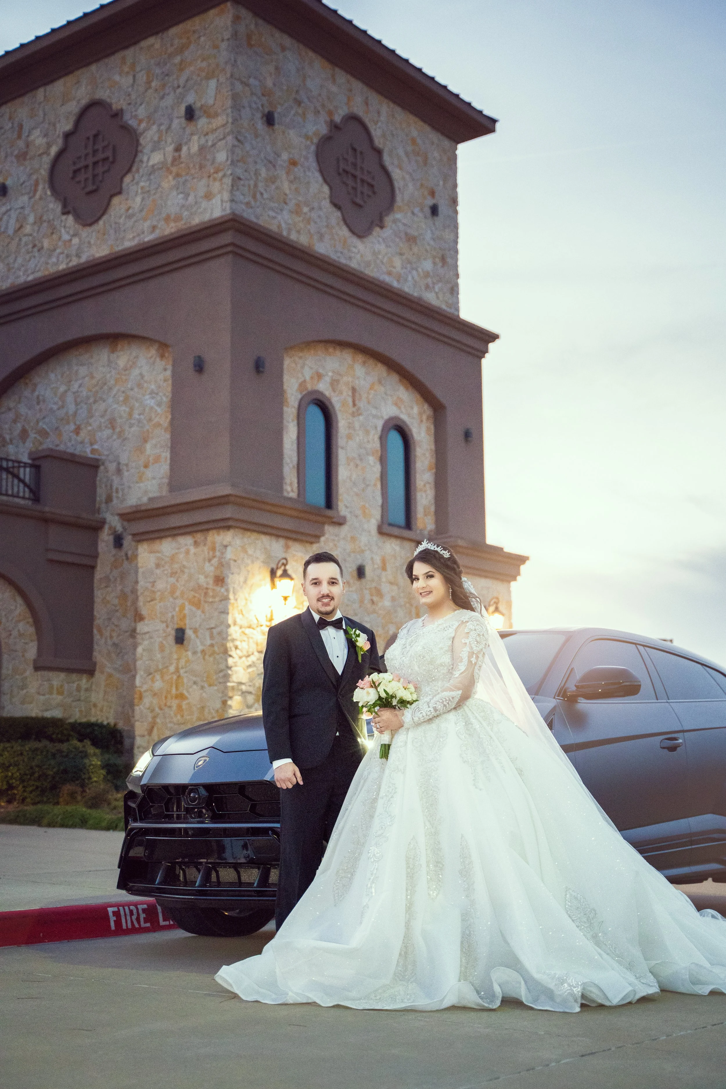 Bride and groom posing in front of a luxury car and building on their wedding day, bride in white gown, groom in tuxedo.