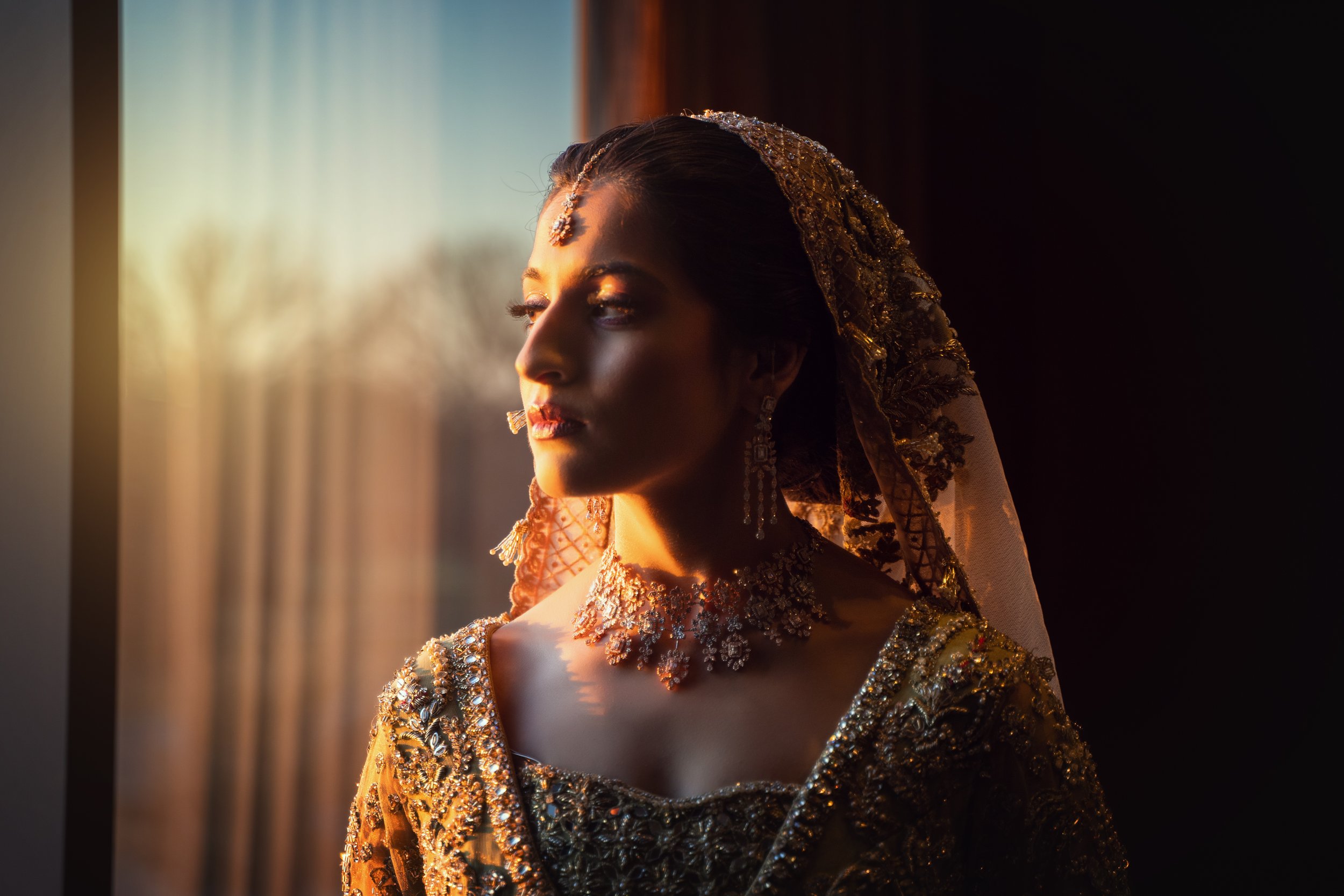 Woman in ornate traditional attire and jewelry looking out a window with soft sunlight.