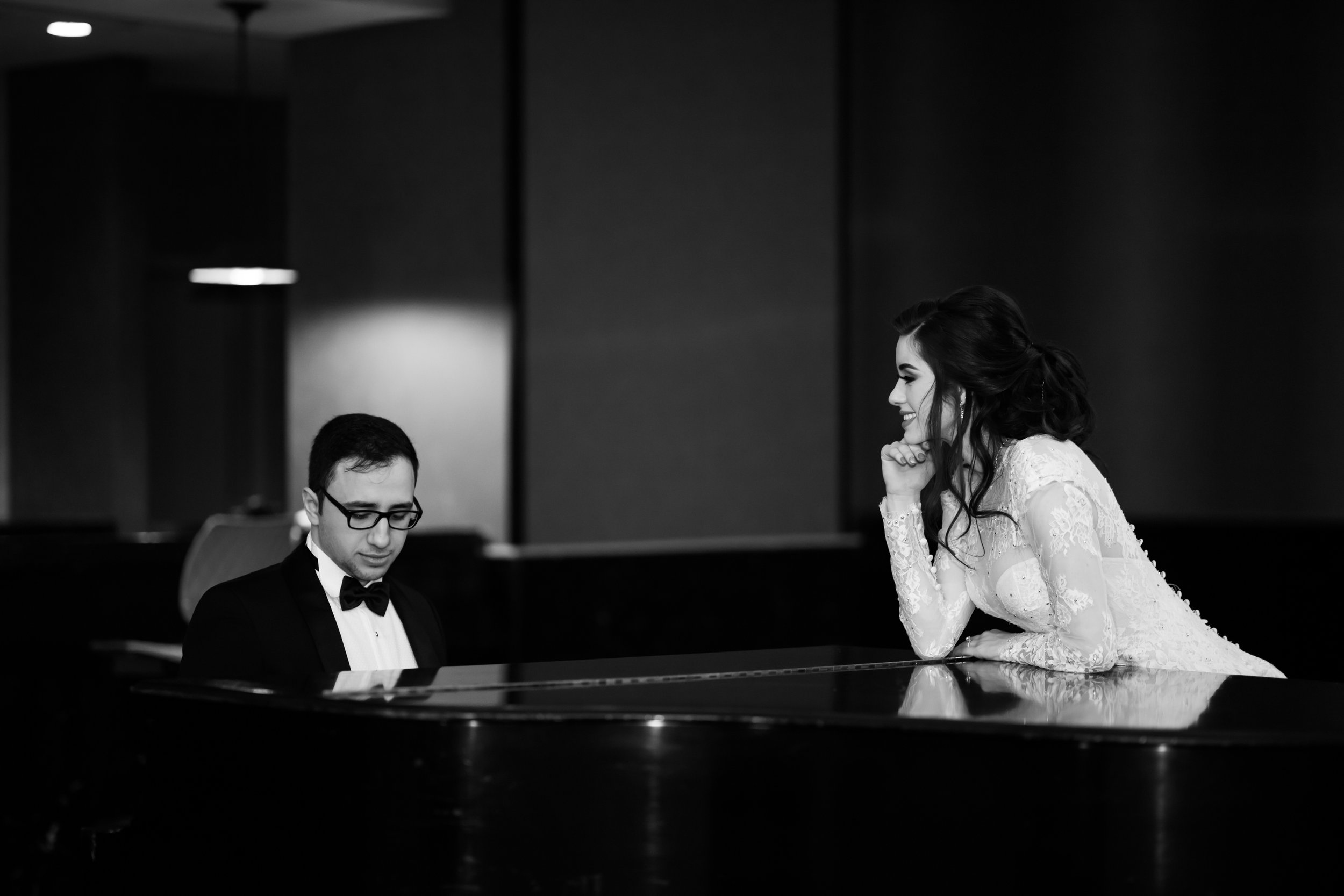 Black and white photo of a man in a tuxedo playing piano, while a woman in a wedding dress leans on the piano smiling at him.