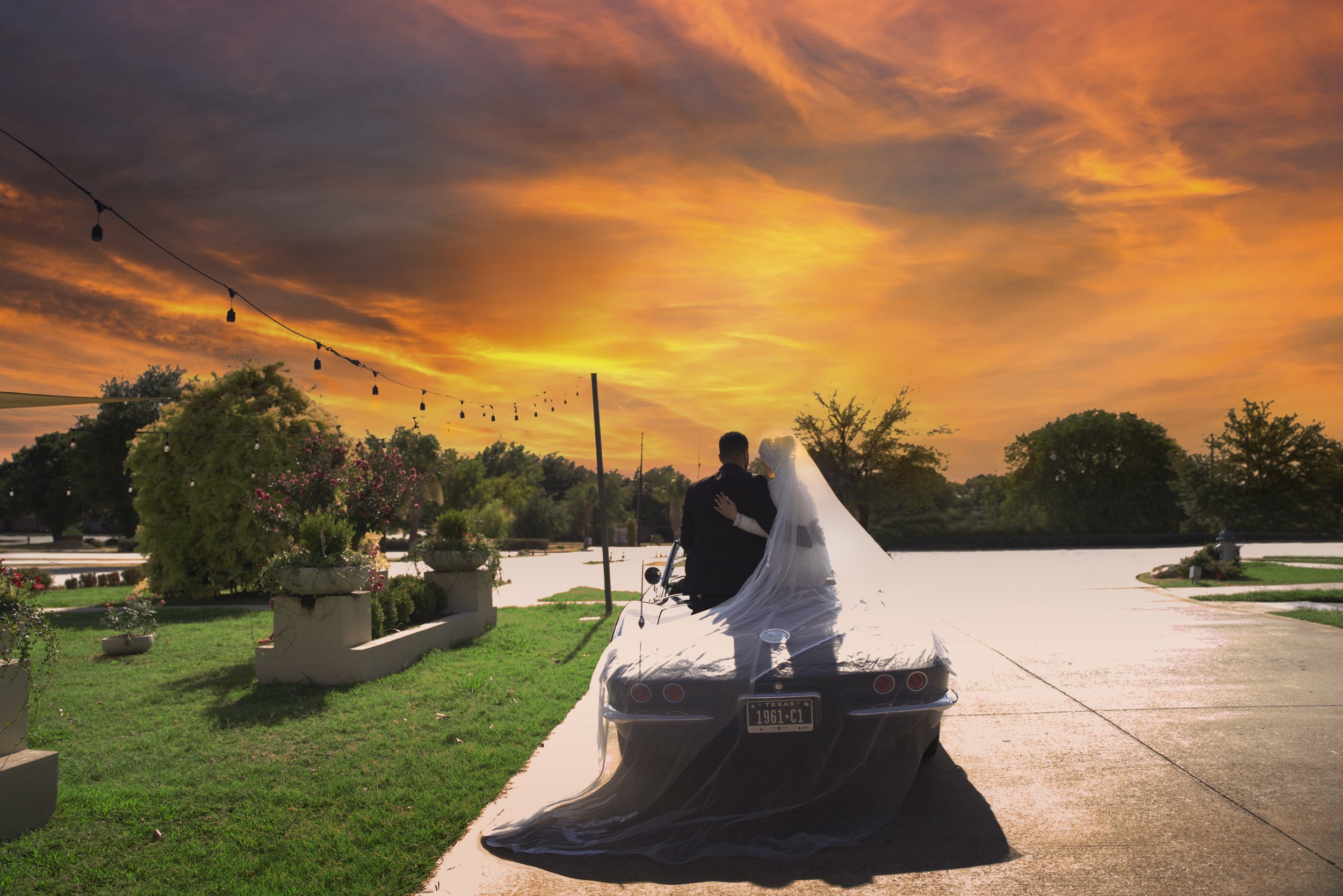 Bride and groom embracing on a car at sunset, with a flowing veil and string lights above, set in a scenic outdoor location.