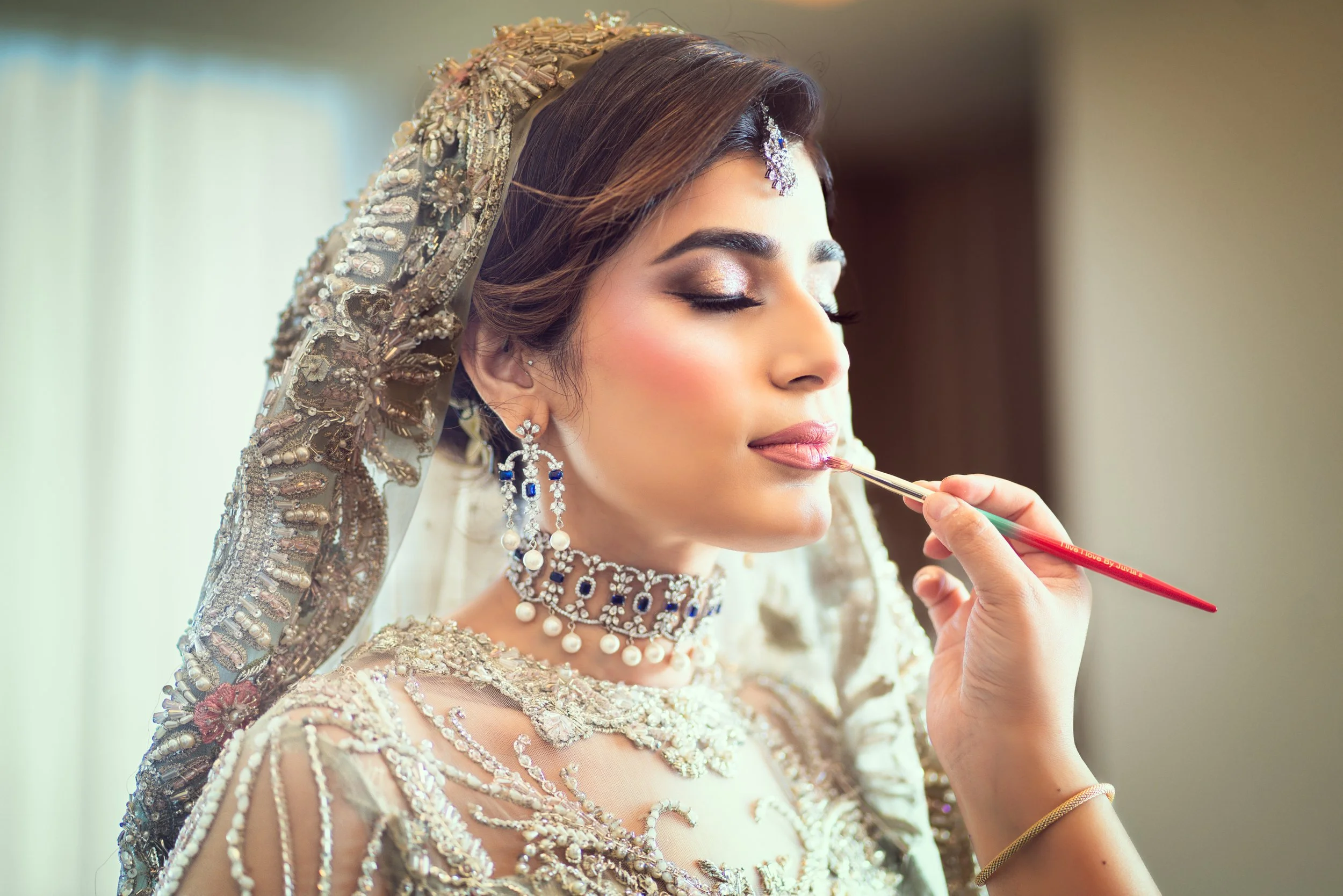 Bride getting makeup applied, wearing an ornate dress with jewelry.