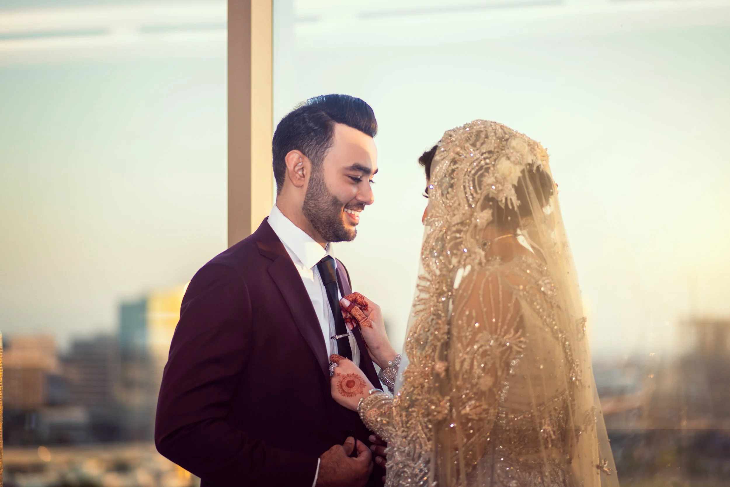 Bride and groom in wedding attire smiling at each other, with the bride adjusting the groom’s tie, against a cityscape backdrop.
