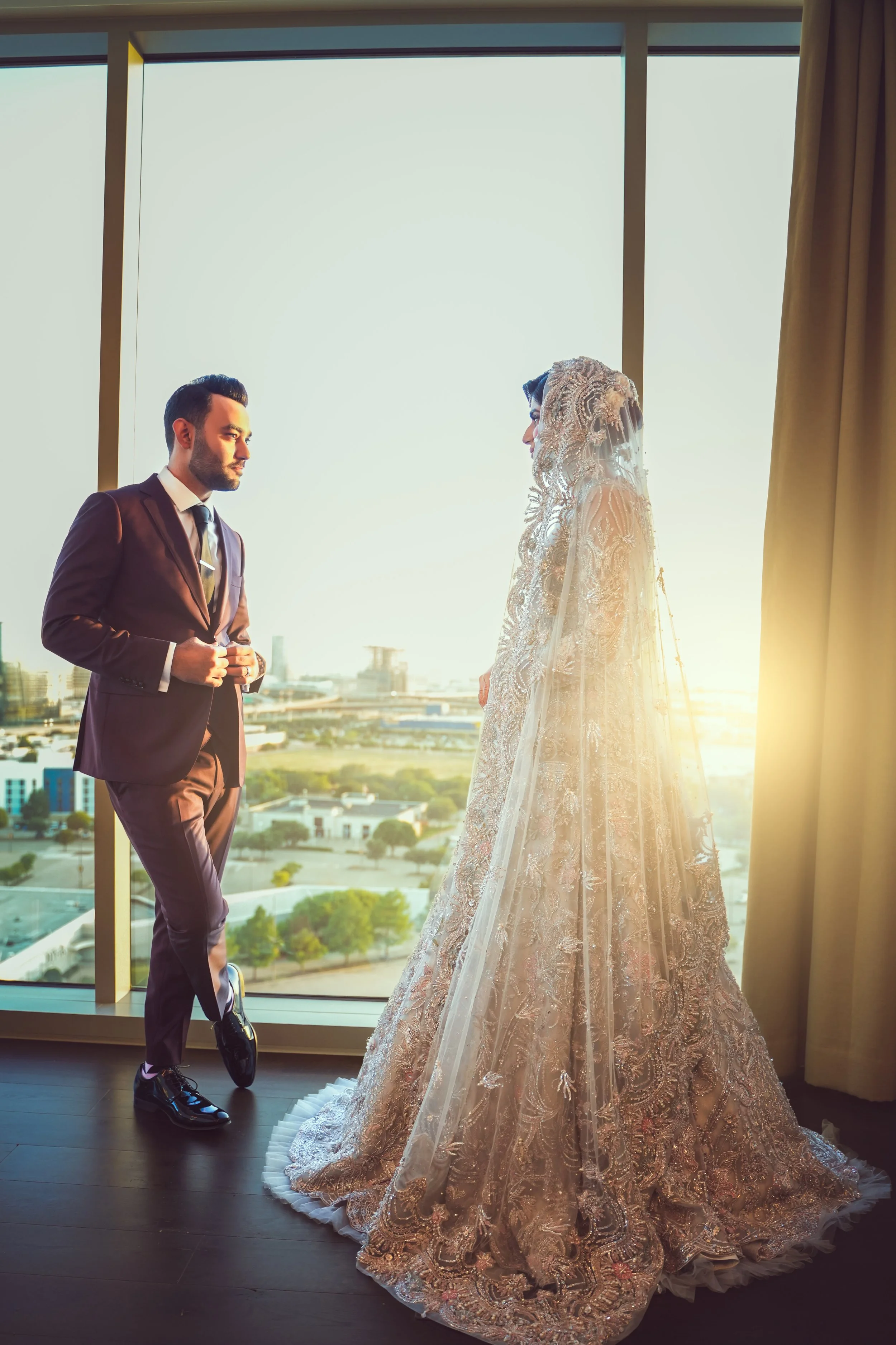 Bride and groom dressed in wedding attire, standing in front of a large window with a cityscape view.