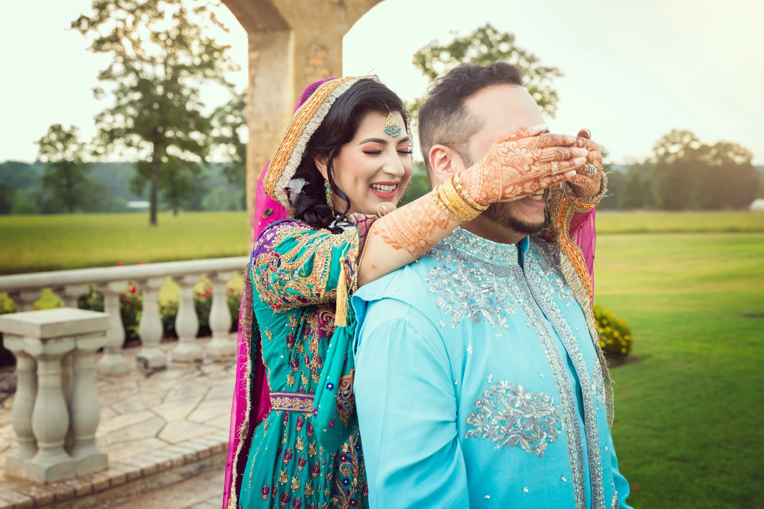 A woman in colorful traditional clothing is playfully covering a man's eyes from behind. Both are outdoors in a landscaped area with greenery and stone railings. The woman has henna on her hands and is smiling.