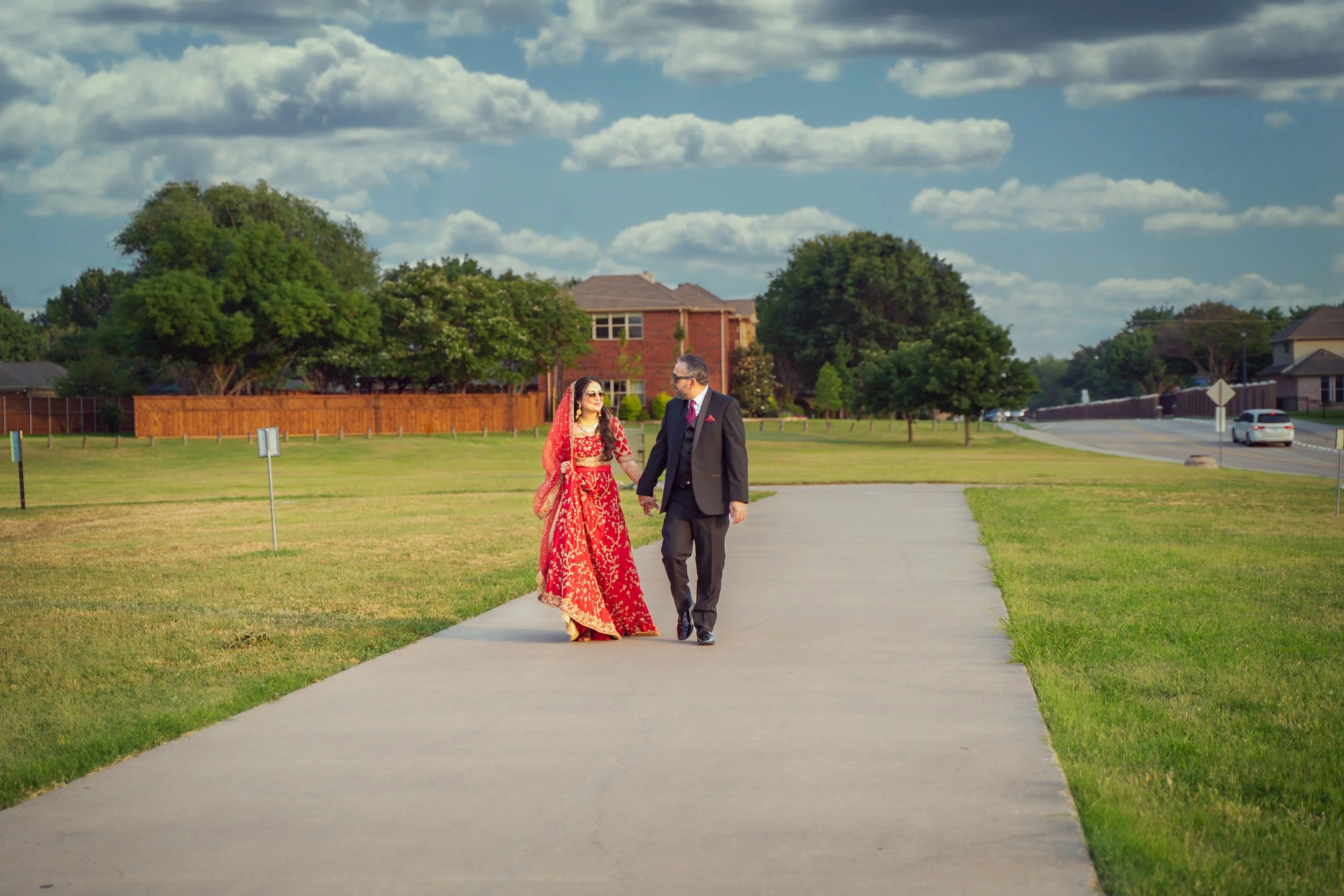 A couple walking on a paved path in a park, with the woman wearing a red traditional outfit and the man in a suit. The background features grassy fields, trees, houses, and a cloudy sky.