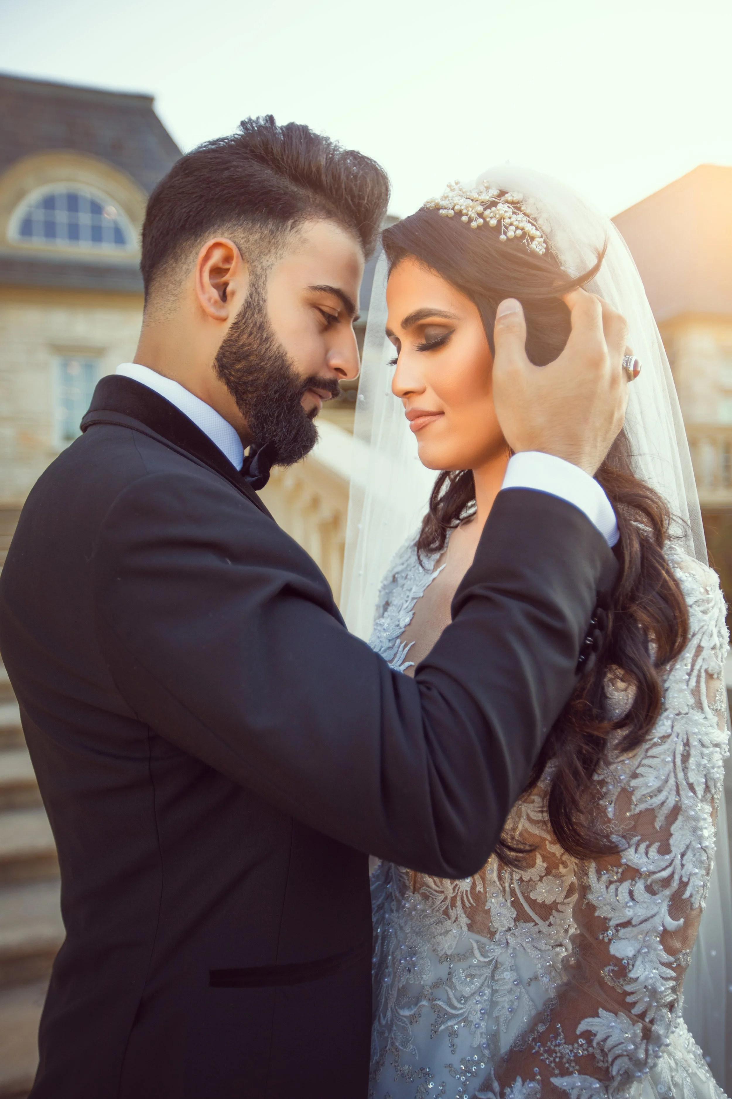 Bride and groom embracing in wedding attire, standing outdoors with soft sunlight.