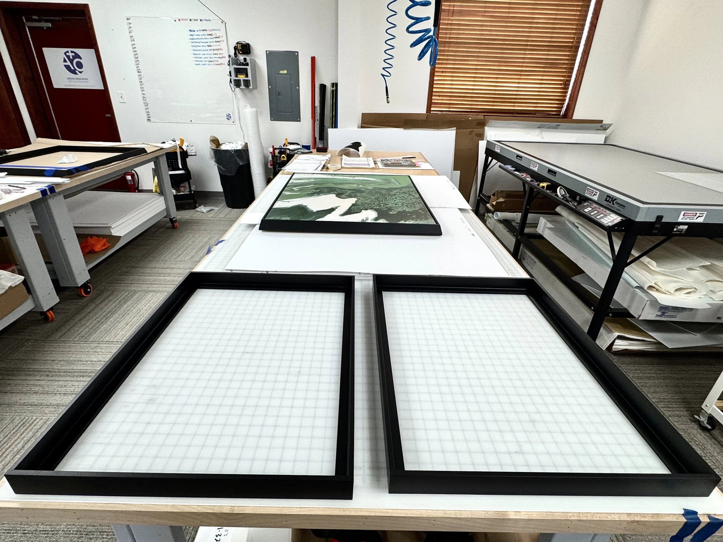 A workspace with several large framed art prints laid out on white tables, surrounded by art supplies, tools, and packaging. A window with closed blinds is in the background.