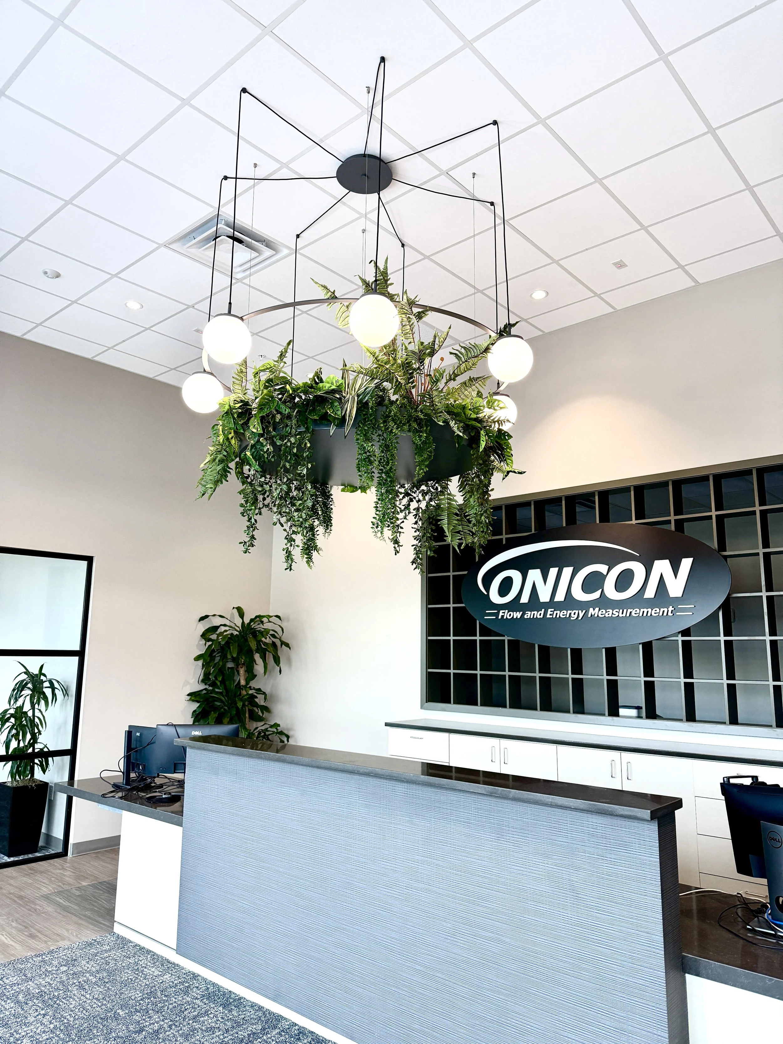 Reception desk in an office with a monitor, potted plants, and a large hanging indoor garden with lights above. A sign reading 'ONICON' and 'Flow and Energy Measurement' is on the wall behind the desk.