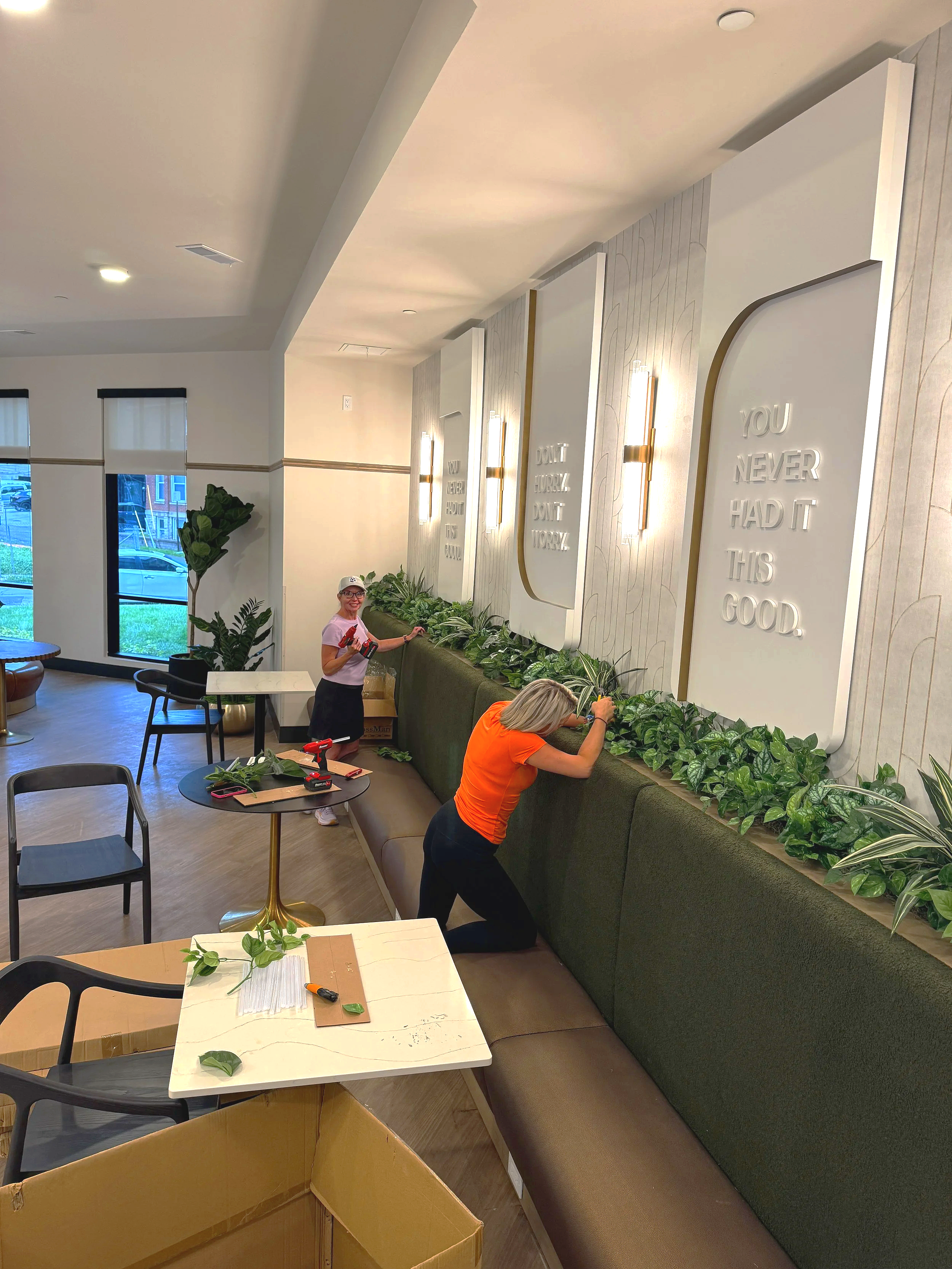 Two women are working on decorating a wall with large white panels that have quote cutouts. One woman in pink is smiling and holding a tool, while the other woman in orange is kneeling and placing green foliage along a bench. The room has large windows, plants, and modern lighting.