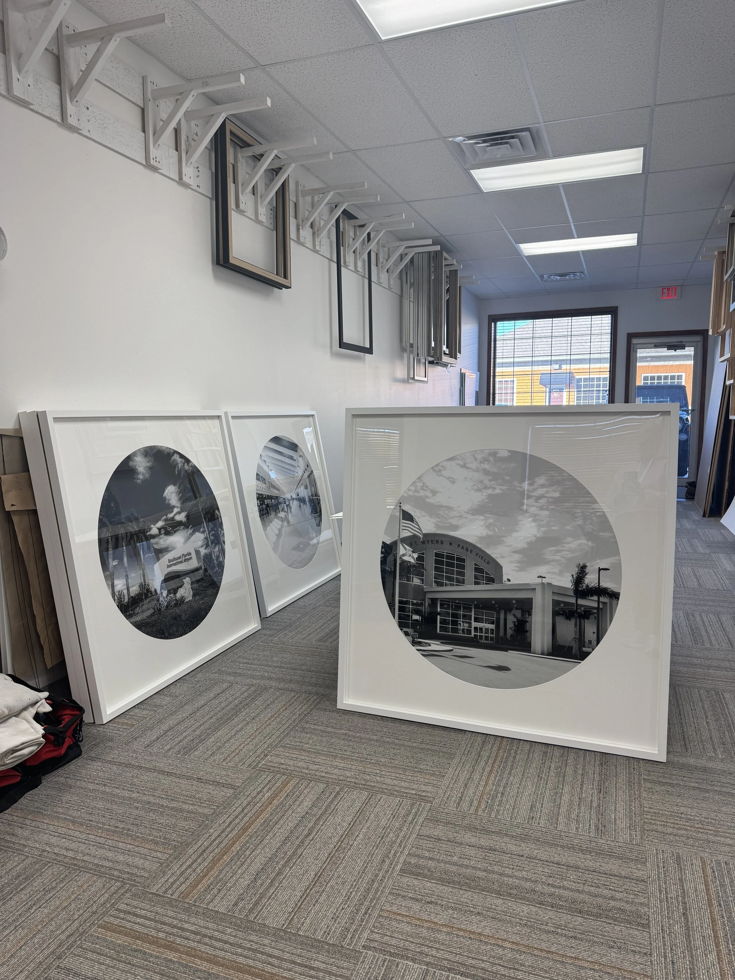Indoor store display with large framed black-and-white photographs of architectural buildings leaning against the wall, and additional frames hanging on the wall, with a window showing a building outside.