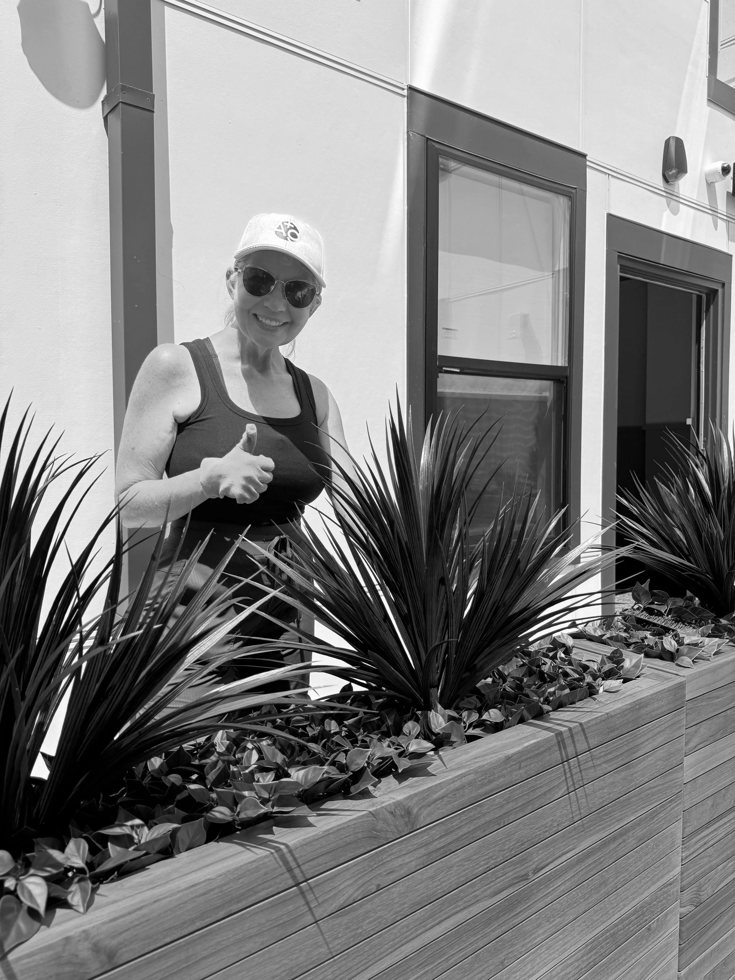 Woman wearing sunglasses and baseball cap smiling and pointing, standing behind a wooden planter filled with tall plants and leafy ground cover in front of modern building windows.