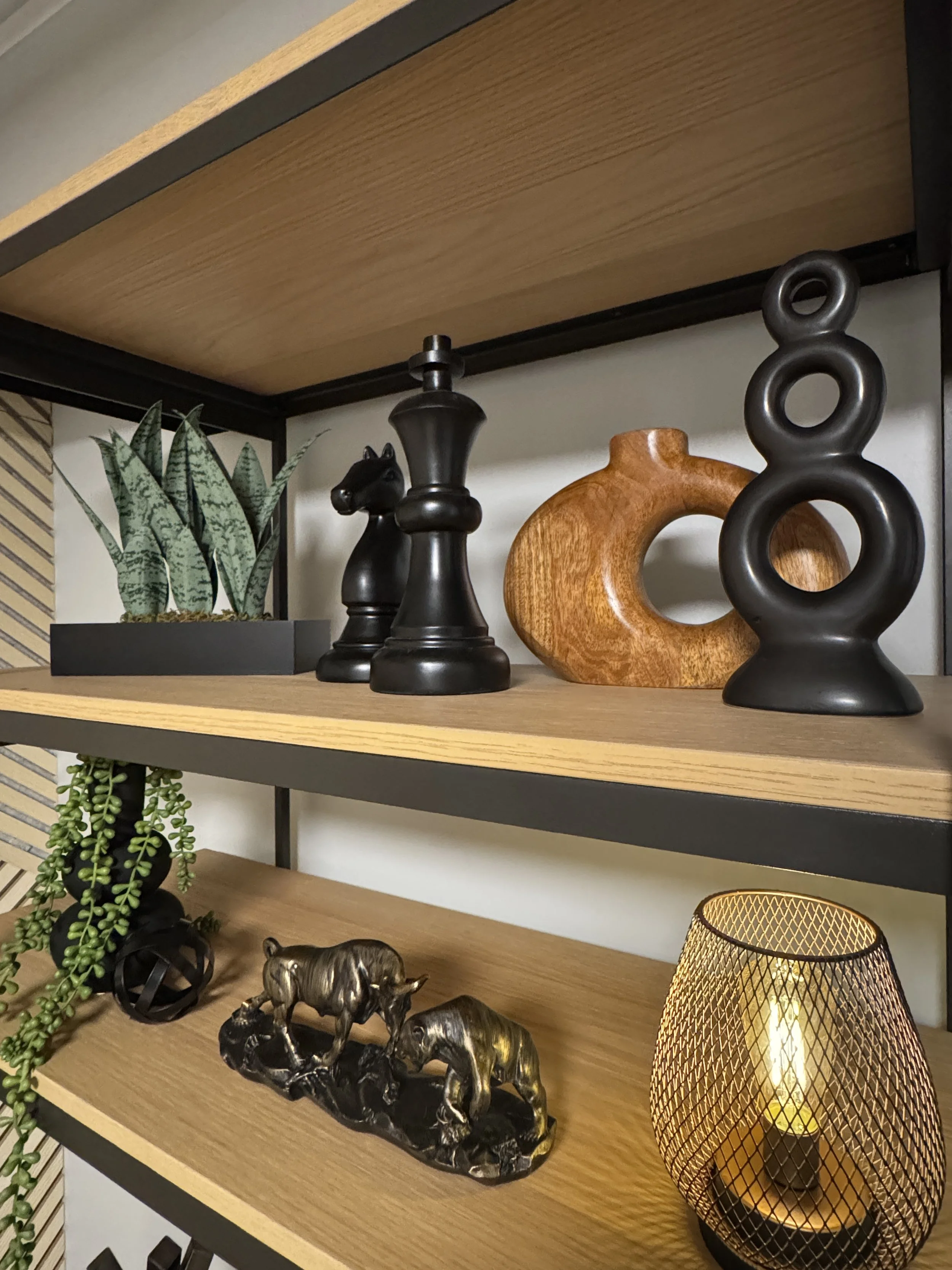 Decorative items on a wooden shelf including potted succulent, black cat figurine, intertwined black sphere, wooden abstract sculpture, black metal sculpture of a rhinoceros, and a lit candle inside a gold wire lantern.