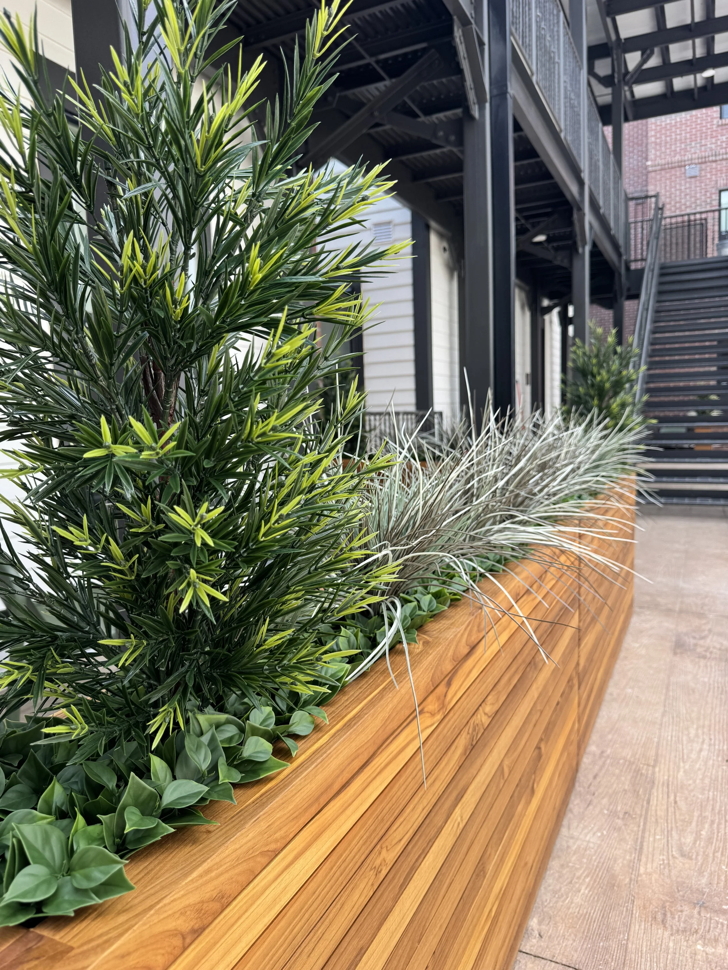 Close-up of a large wooden planter filled with various green and gray plants, situated in a multifamily apartment courtyard outside a building with stairs and black metal railings in the background.