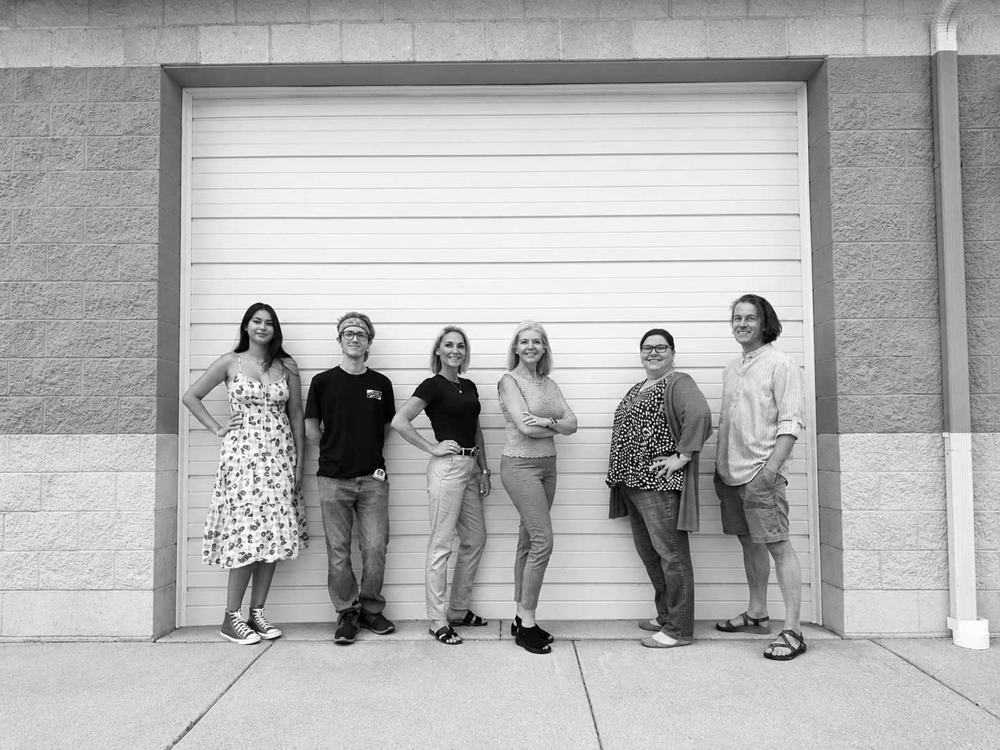 Group of six diverse adults standing in front of a large white garage door, smiling and posing outdoors.