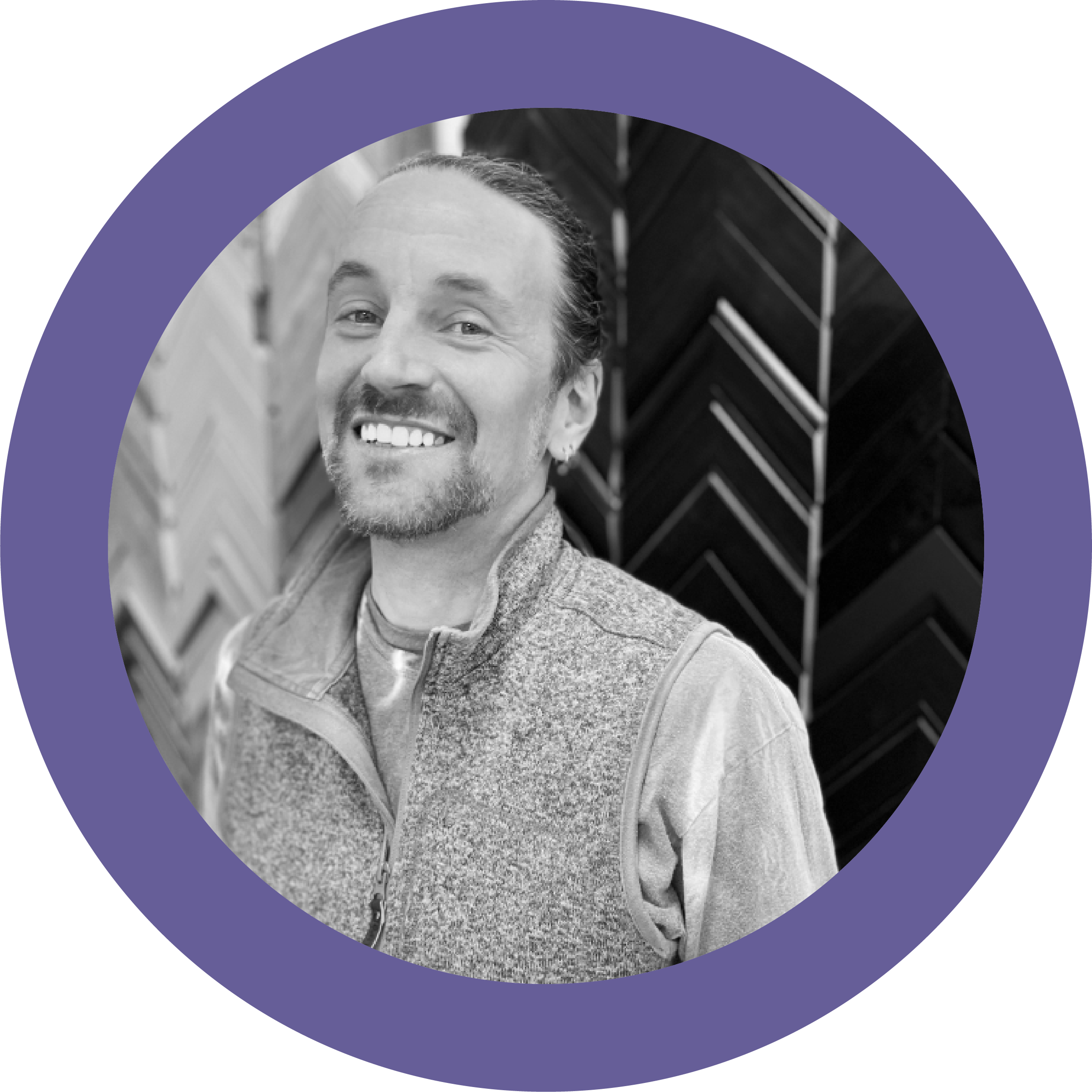 Black and white photo of a man with a big smile, short hair, and a beard, wearing a light-colored jacket over a shirt, standing in front of a textured wall with geometric patterns.