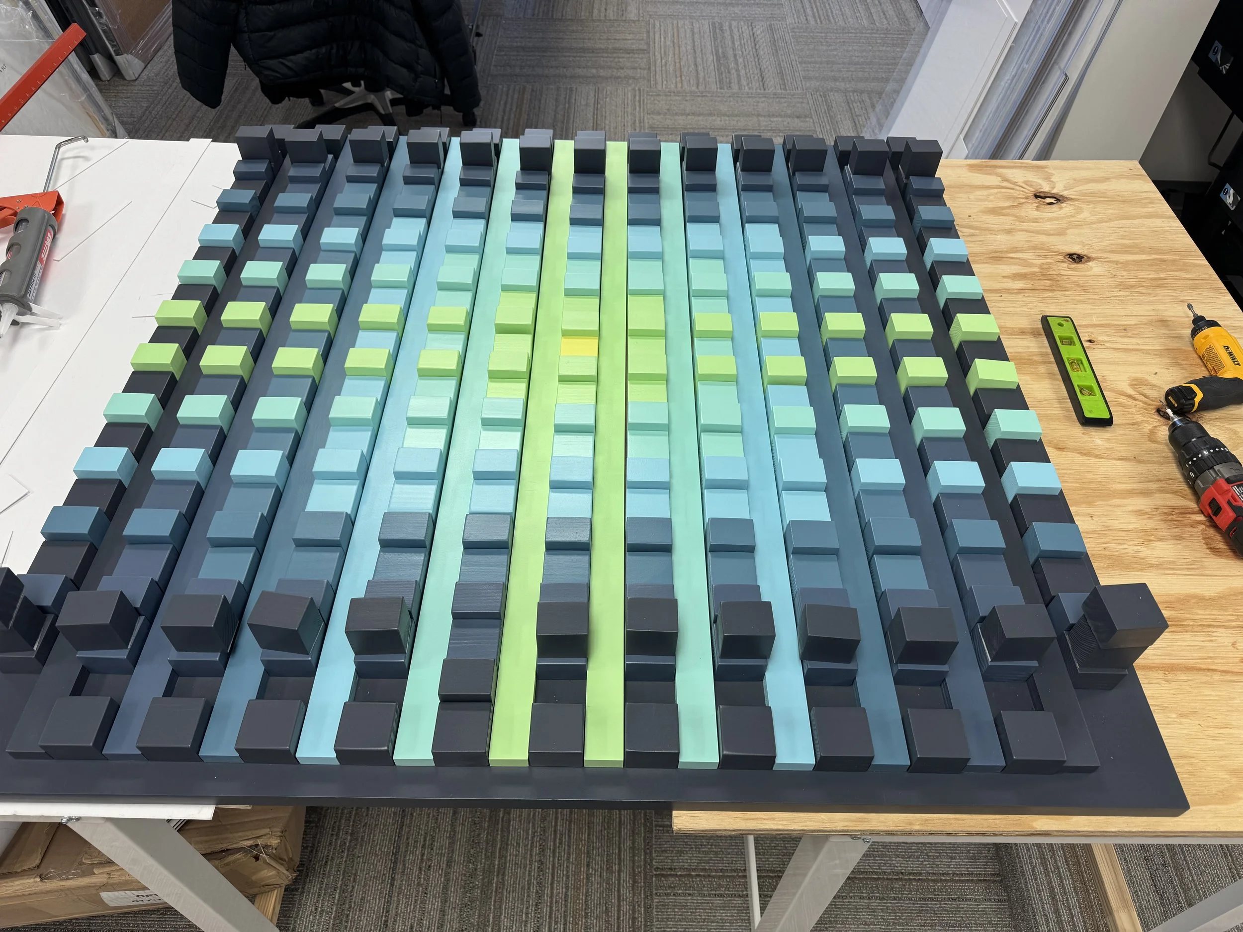 A tabletop with organized foam blocks in shades of blue, green, and black, along with tools including a cordless drill, a spirit level, a glue gun, and a small screwdriver, on a wooden surface in a workshop or office environment.