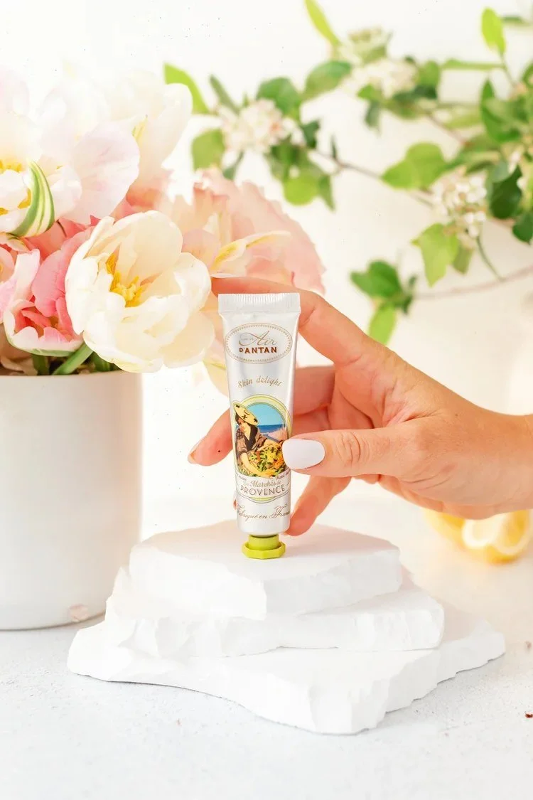 A hand holds a skincare tube among pink peonies, citrus, and white blocks.