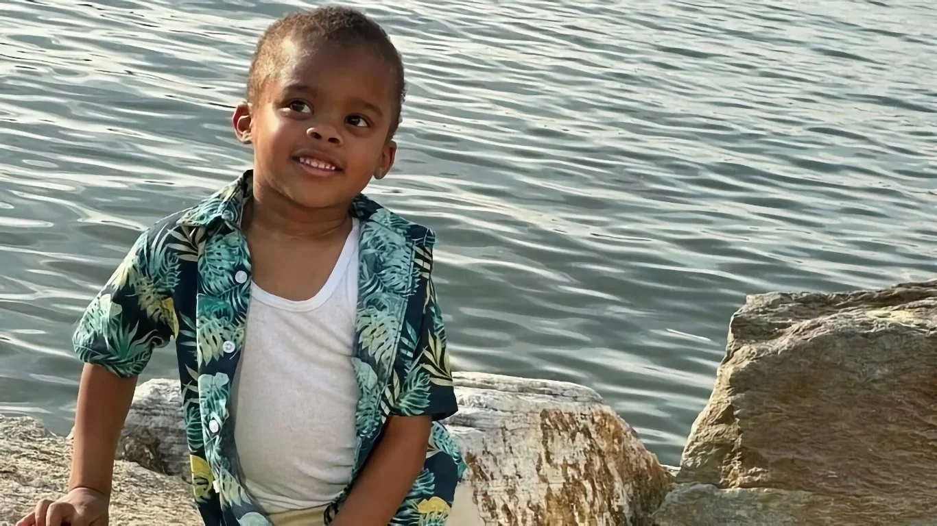 A little boy in a floral shirt sits on boulders next to a body of water.
