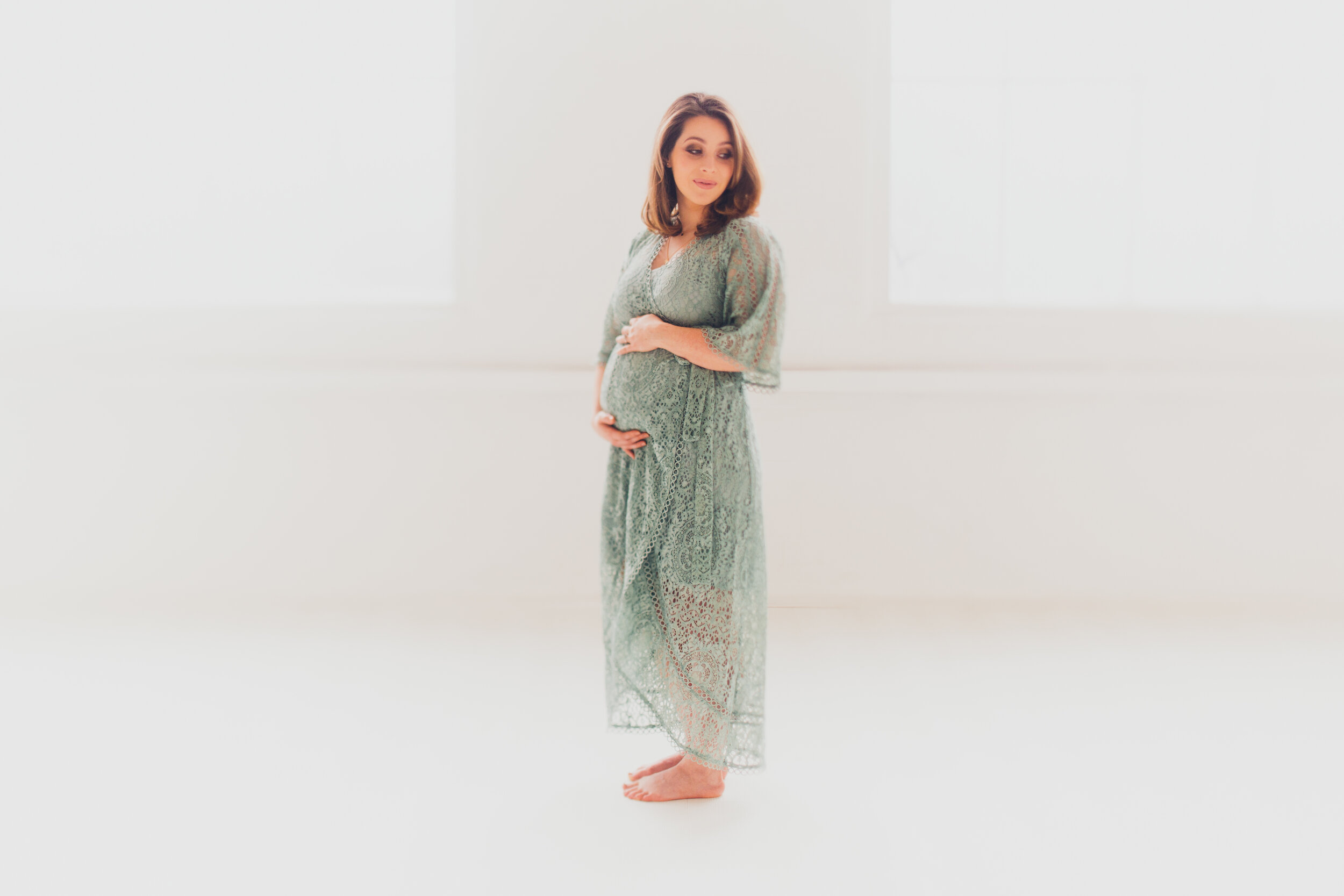 Pregnant woman standing in a soft white photography studio, cradling her baby bump