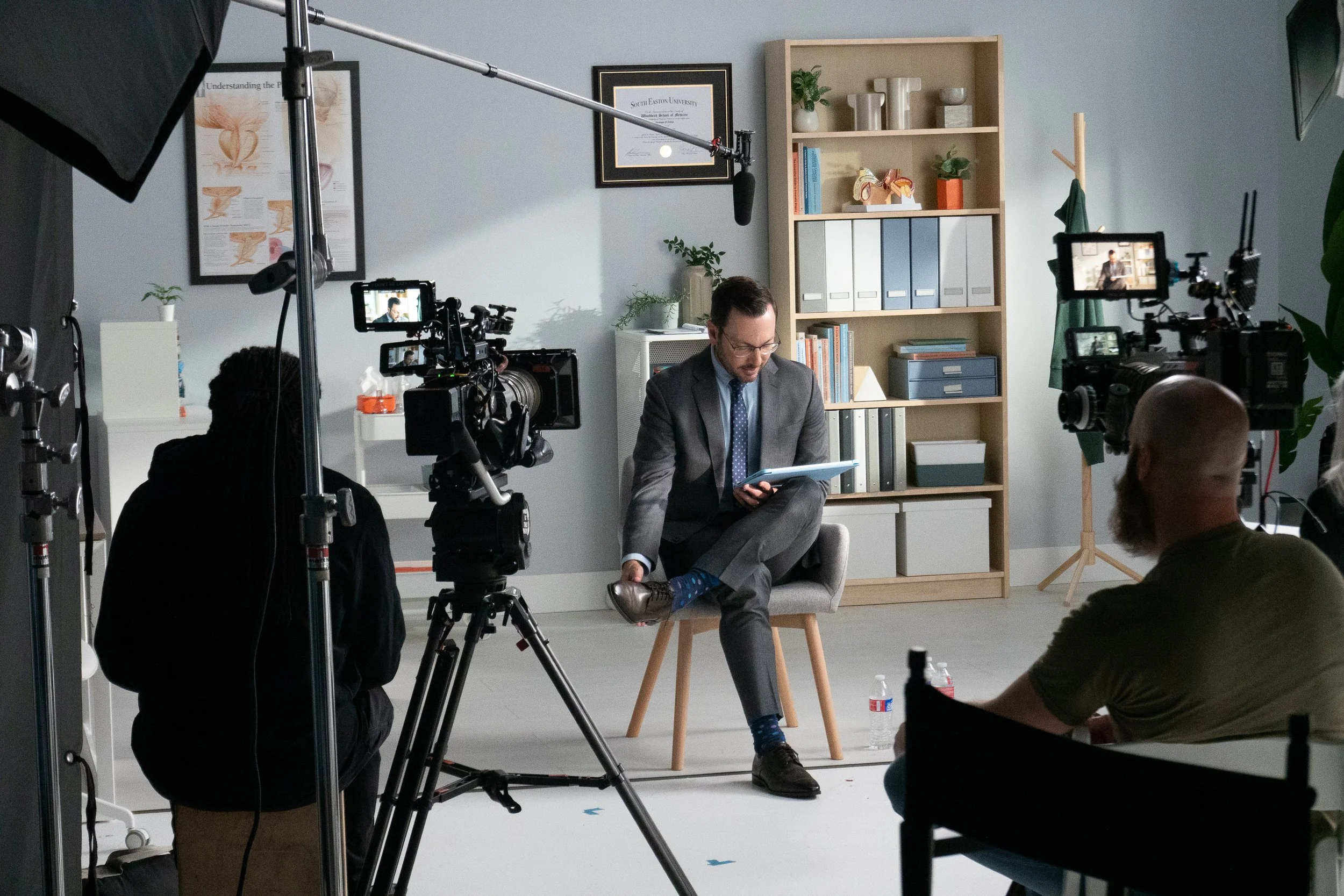 A man in a suit and blue shirt with a tie, seated on a chair, being filmed in a recording studio or office set with filming equipment and cameras around him.